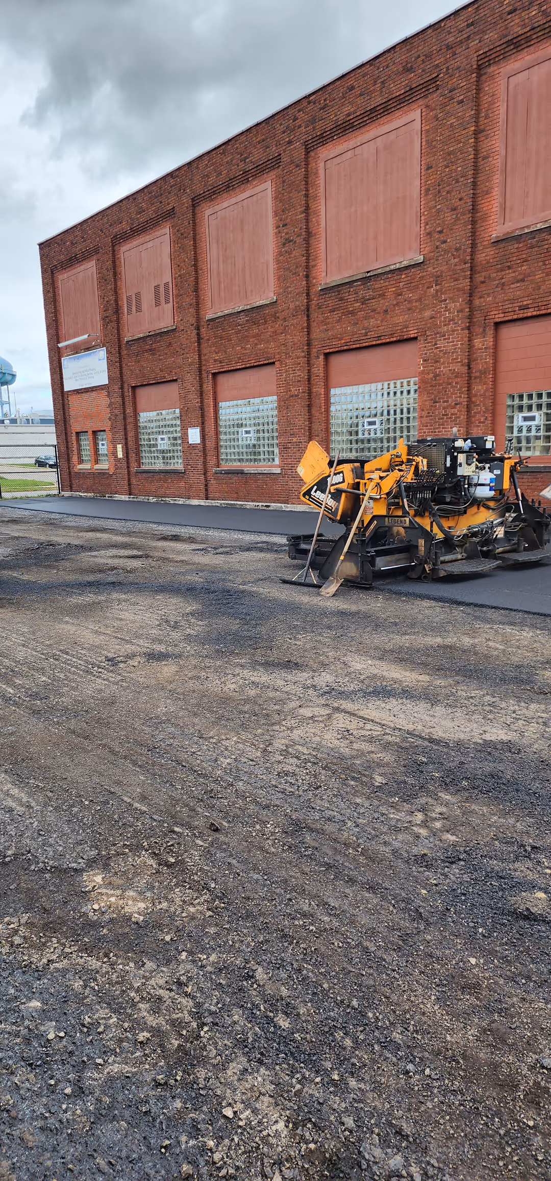 Construction vehicle paving fresh asphalt along the side of a red brick industrial building under a cloudy sky.
