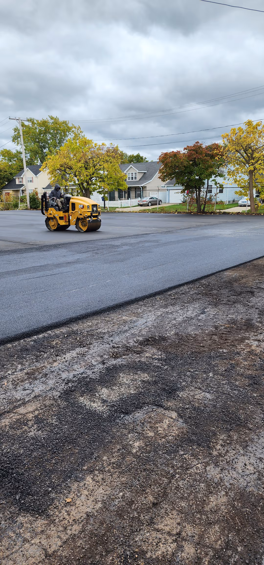 Yellow road roller compacting fresh asphalt on a residential street with houses and trees under a cloudy sky.