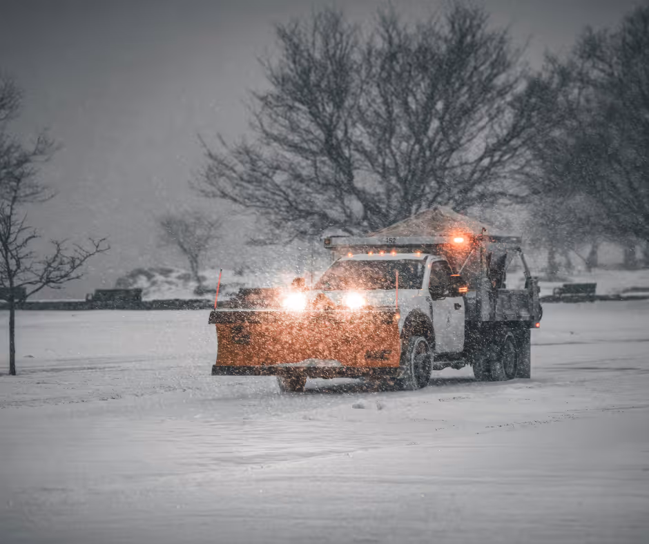 White snow plow truck clearing snow on a snowy road with trees in the background during a snowstorm.