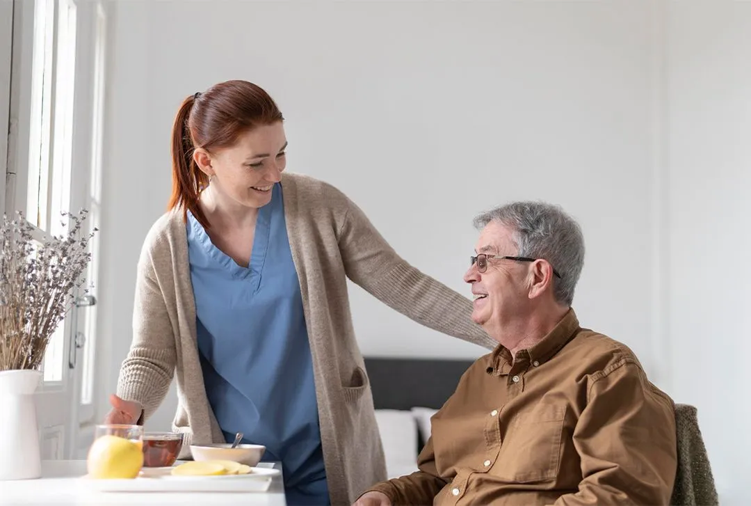 Caregiver in blue scrubs smiling while serving tea to elderly man in brown shirt at table