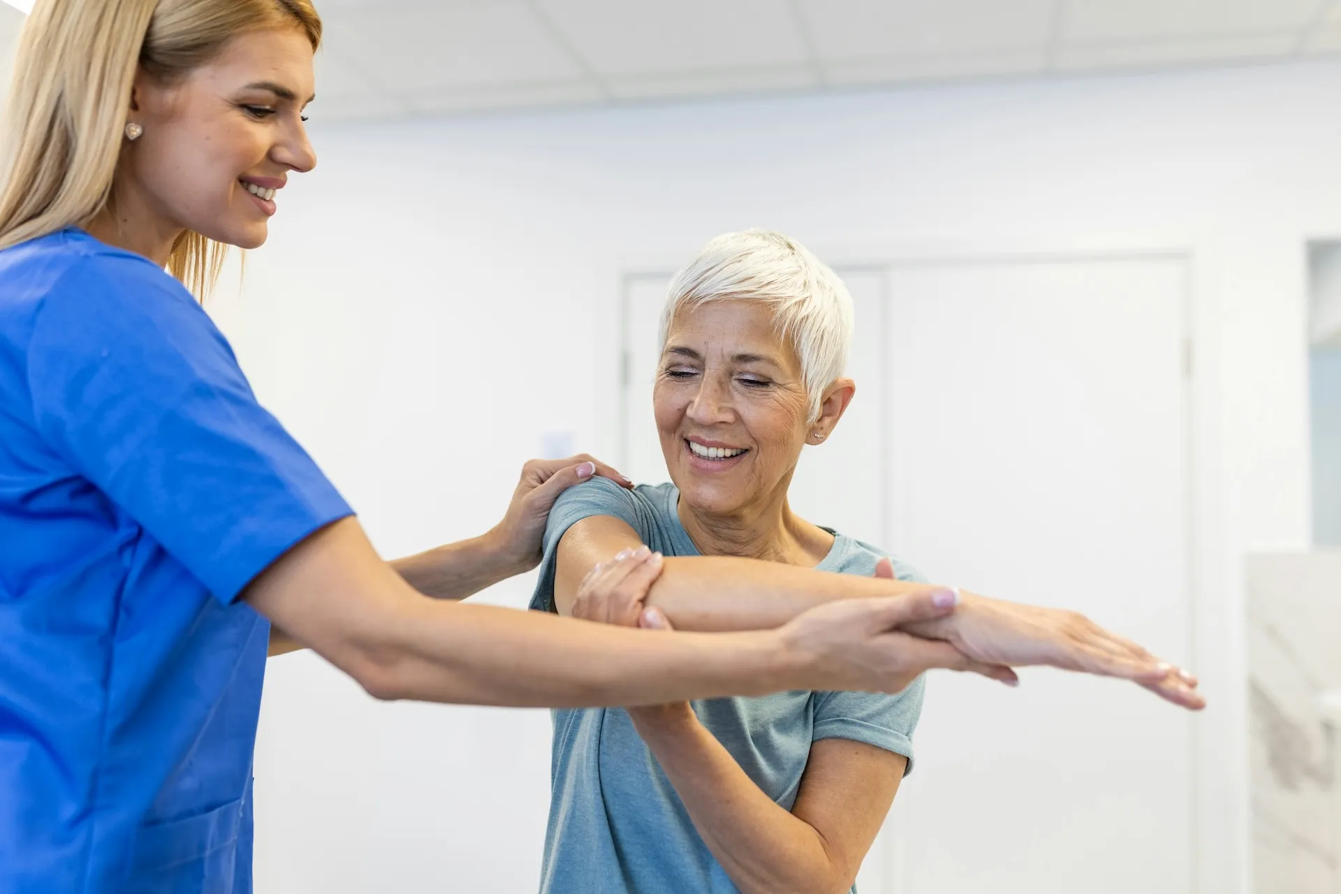Healthcare worker in blue scrubs assists elderly woman with arm exercises in clinic