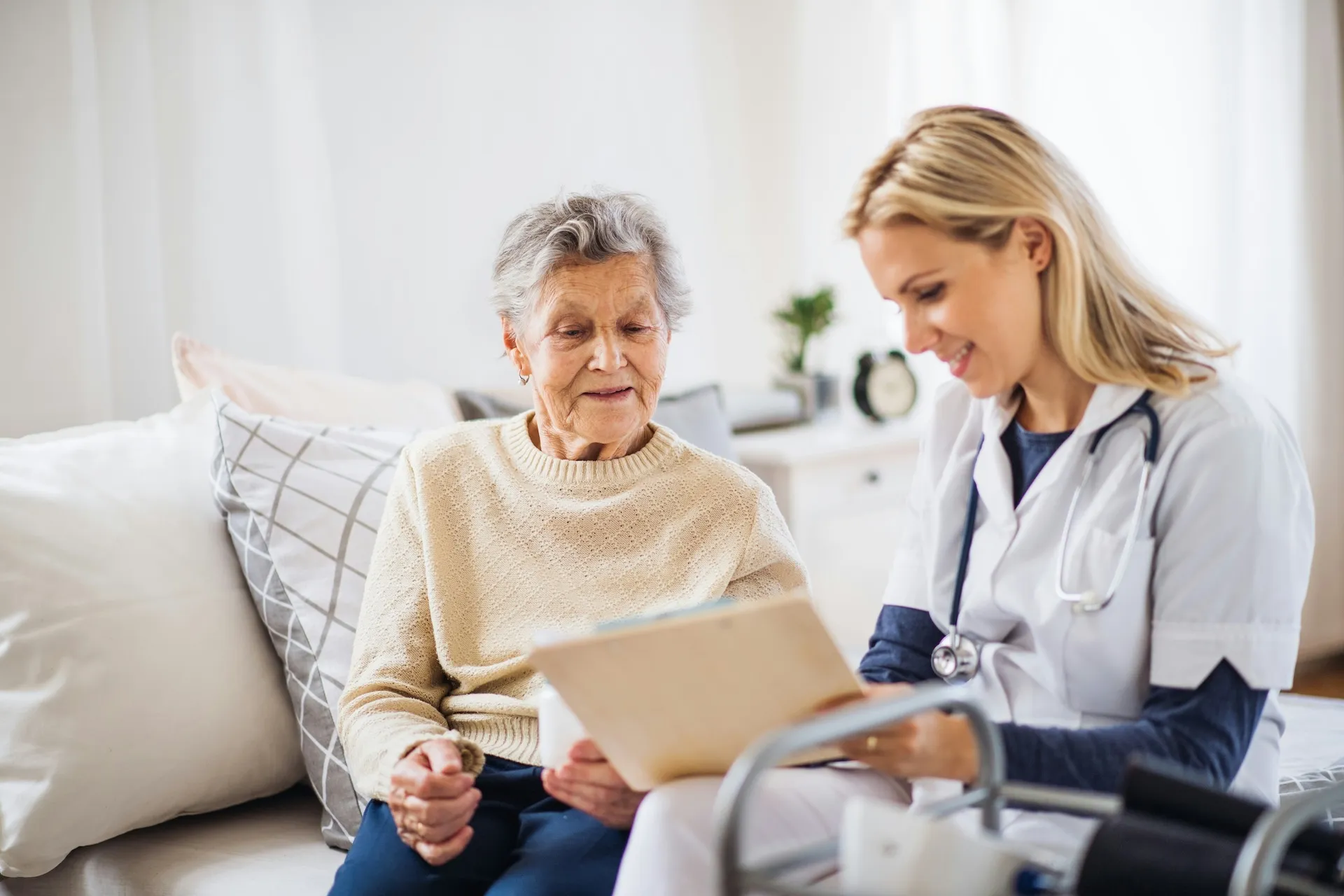 Female doctor with stethoscope reviews tablet with smiling elderly woman patient on couch.
