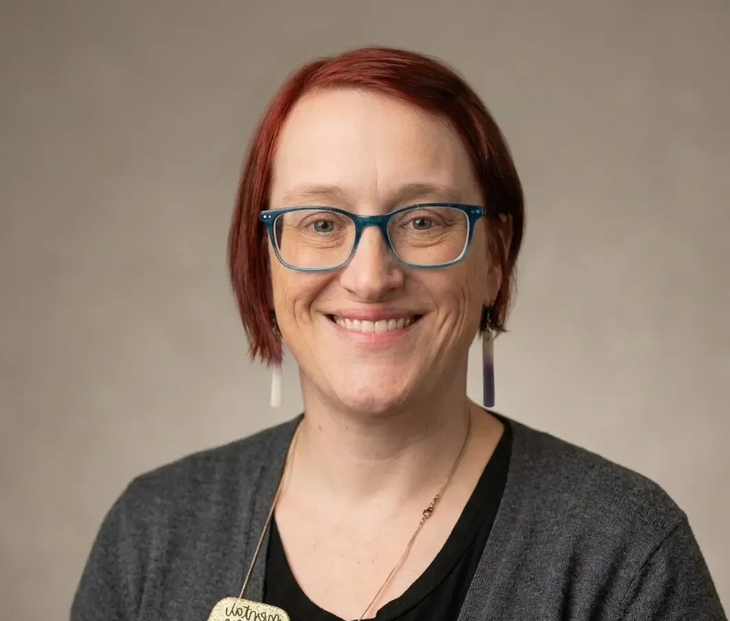 Smiling person with red hair and blue glasses wearing dark gray shirt