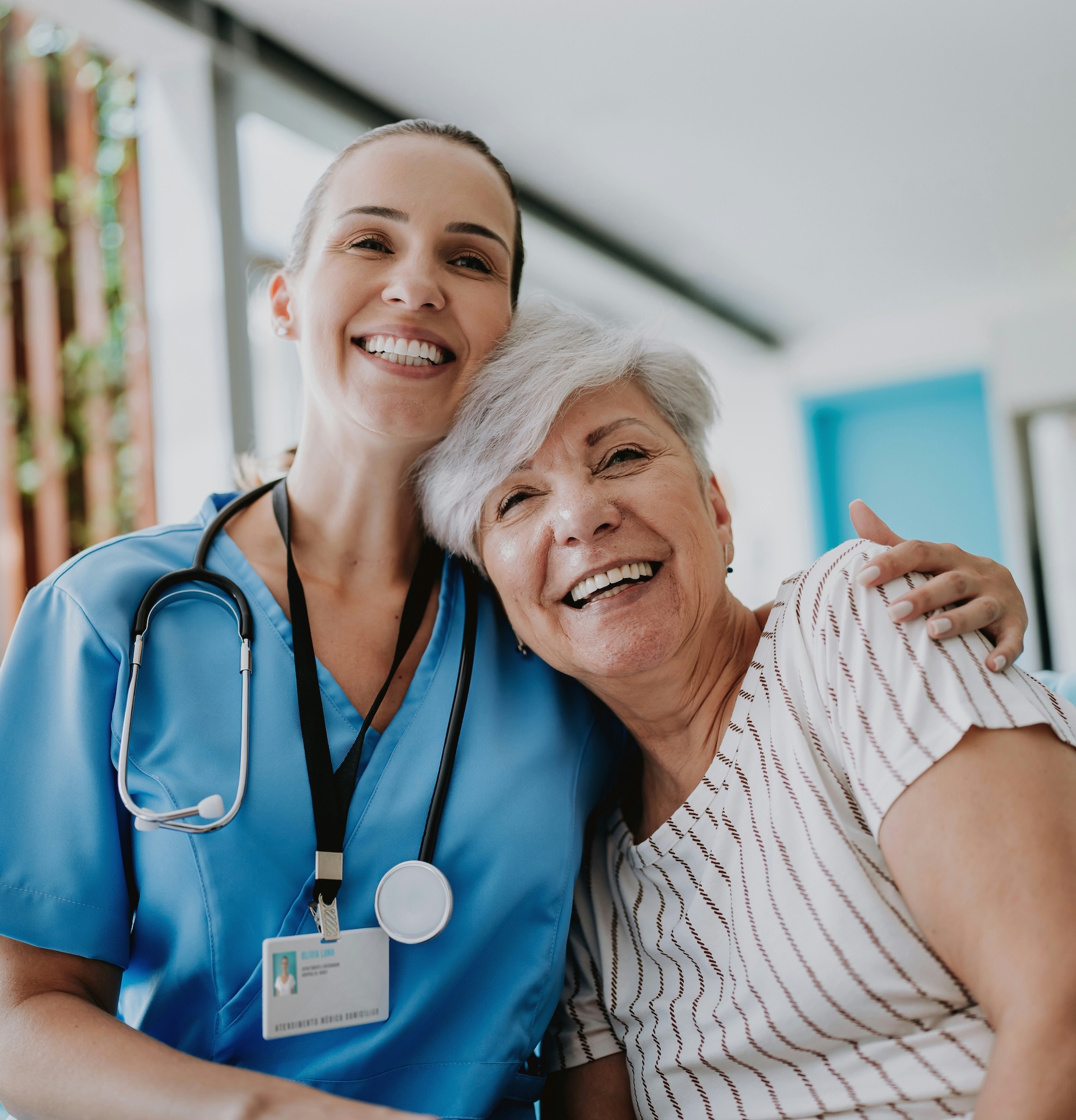 Smiling female nurse in blue scrubs with stethoscope posing with elderly female patient indoors