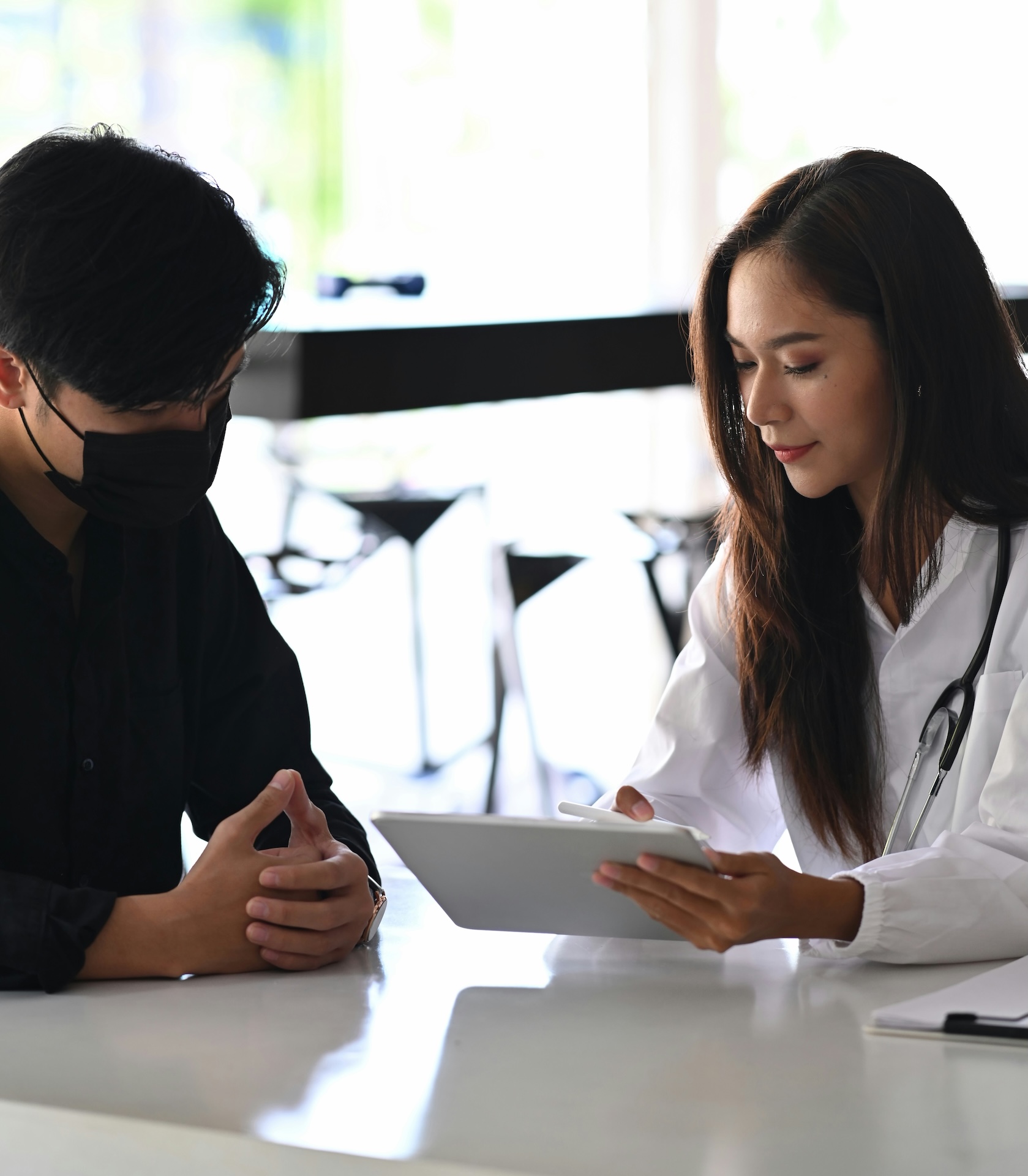 Female doctor showing tablet to masked male patient in modern clinic setting