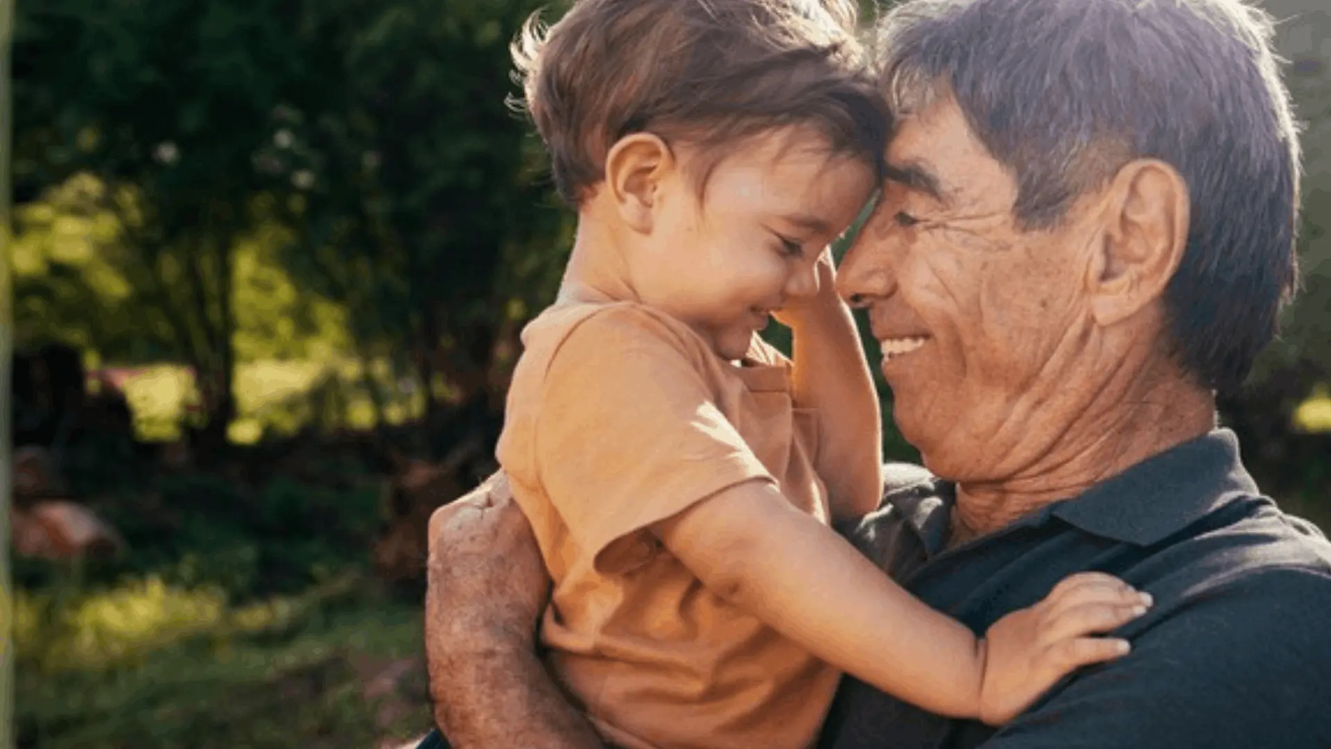 Smiling elderly man holding a young child close, touching foreheads outdoors with green foliage background.