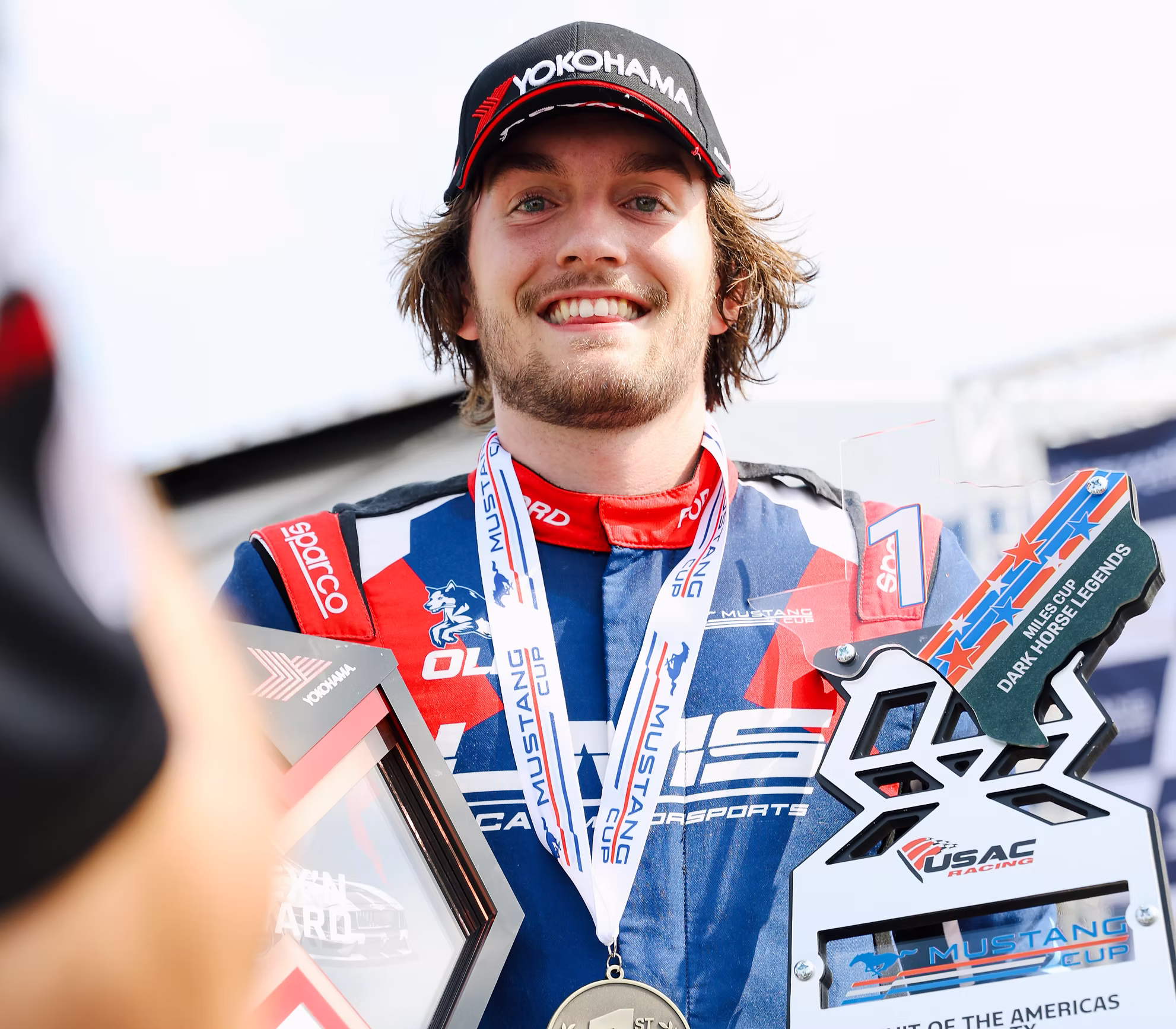 Smiling race car driver in racing suit and Yokohama cap holding Mustang Cup trophy and wearing first place medal.