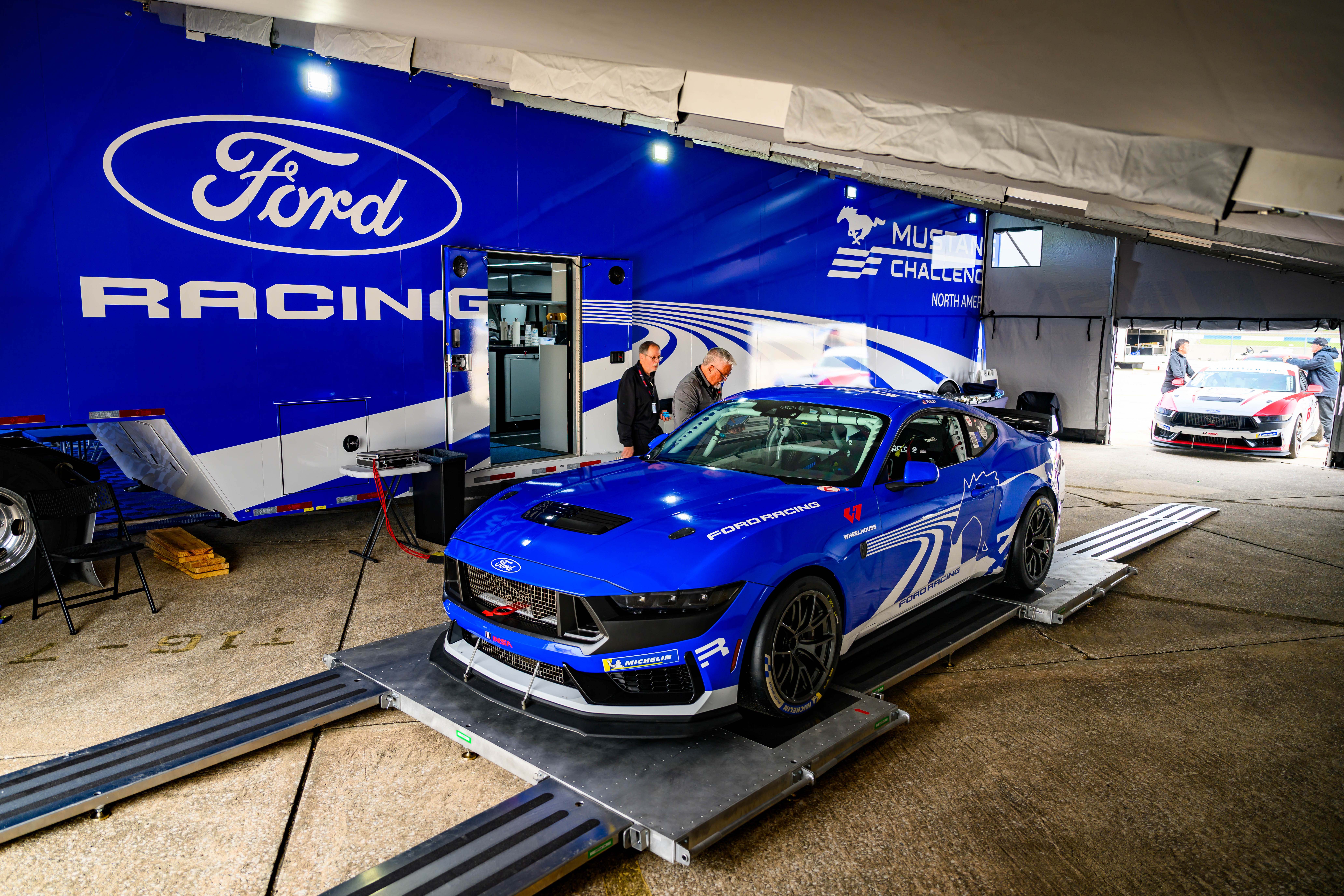 Blue Ford Mustang race car on a platform inside a garage with Ford Racing branding and two people behind it.