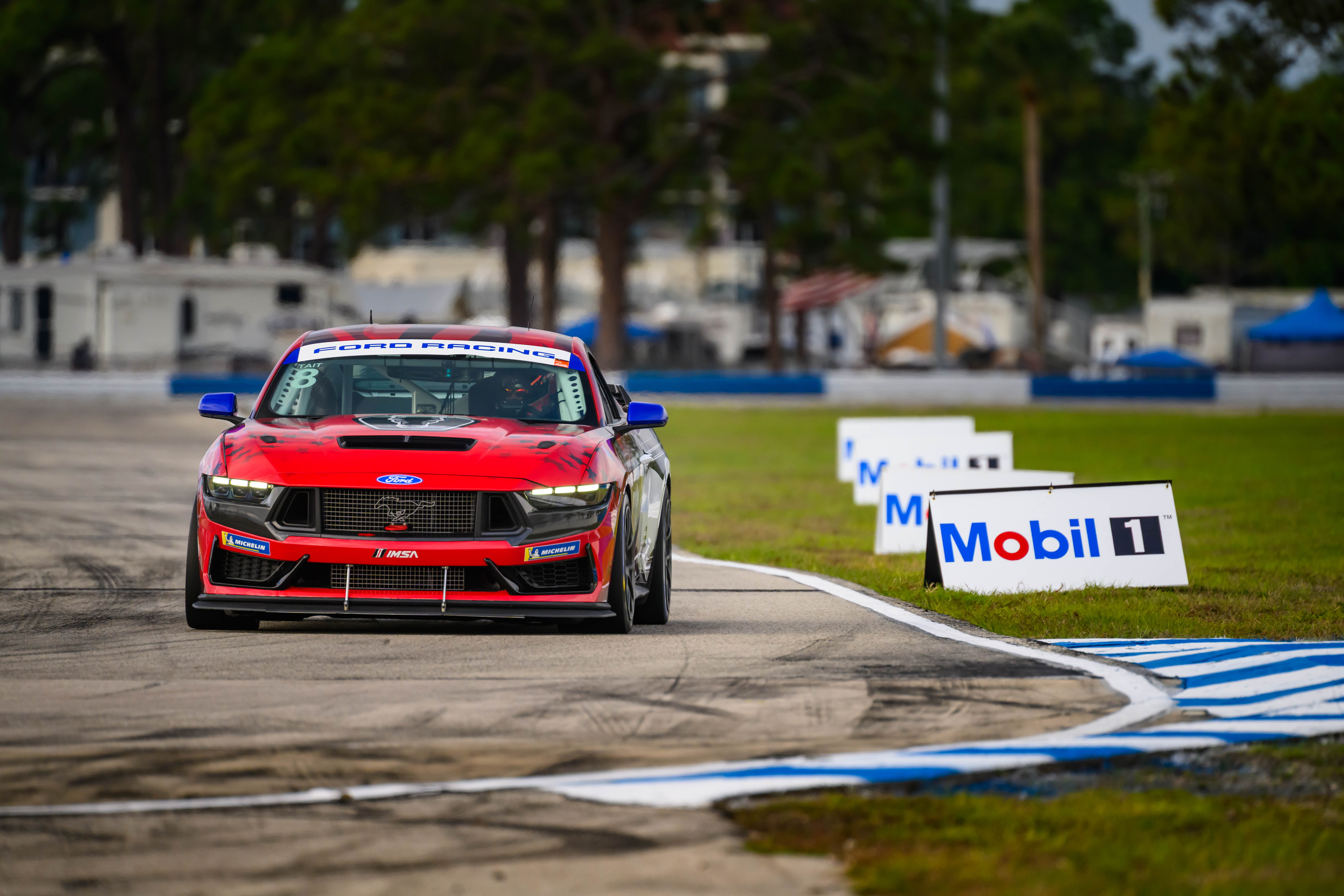 Red Ford Mustang race car on a racetrack next to Mobil 1 sponsor signs.