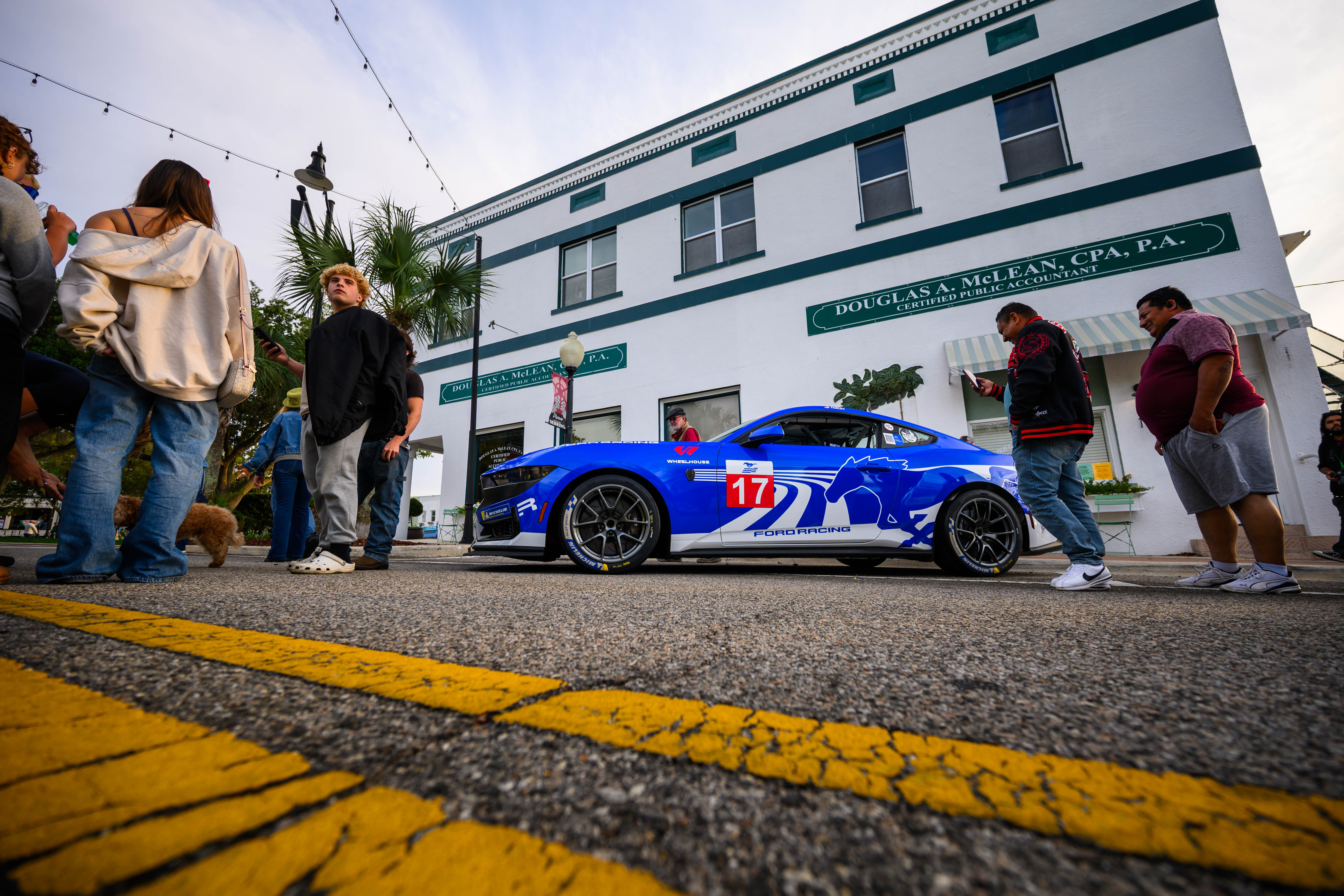Blue Ford Mustang race car with number 17 parked on a street with people and a white building with green trim in the background.