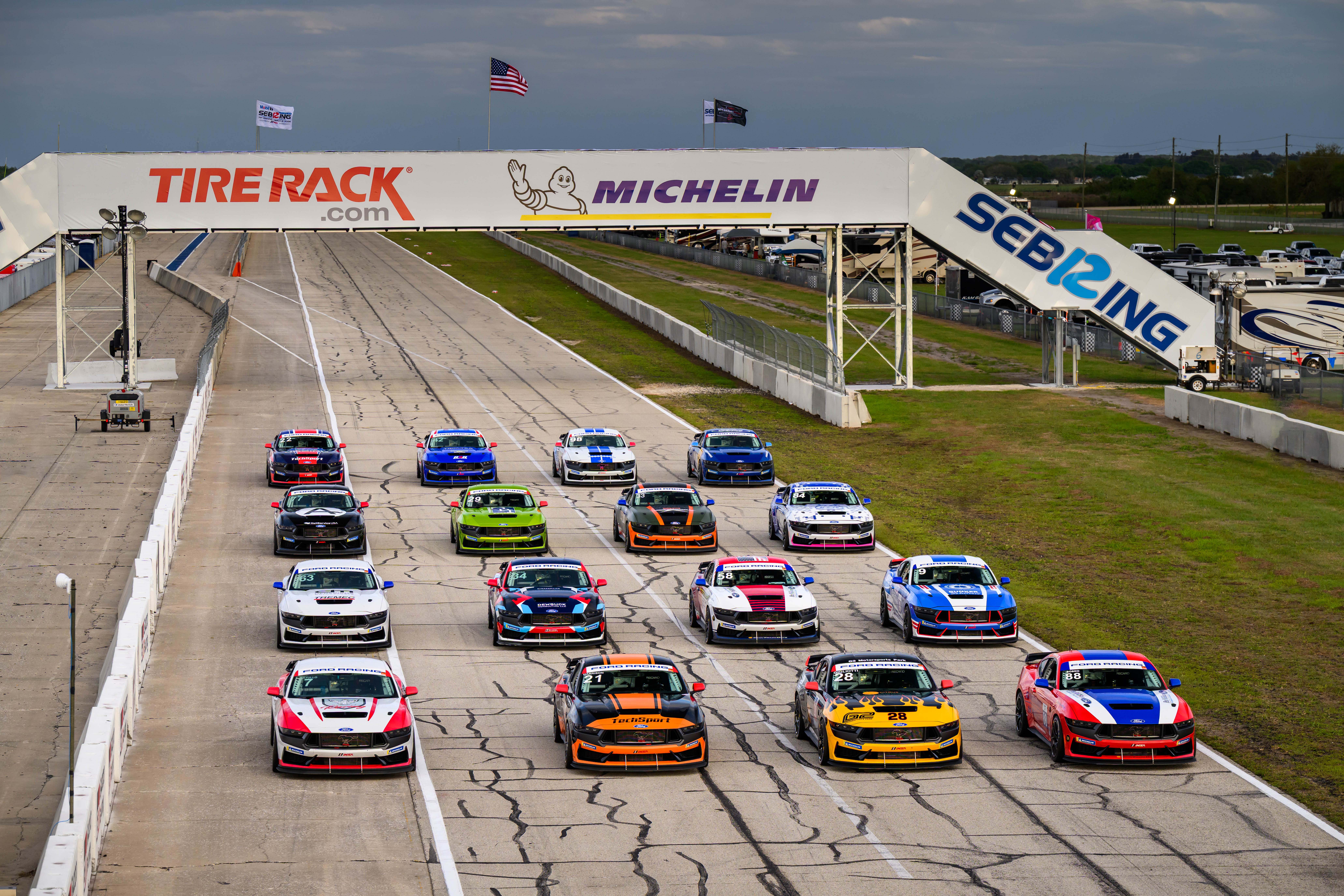 A lineup of colorful race cars positioned on the starting grid of Sebring raceway under an overhead sign with Tire Rack, Michelin, and Sebring logos.