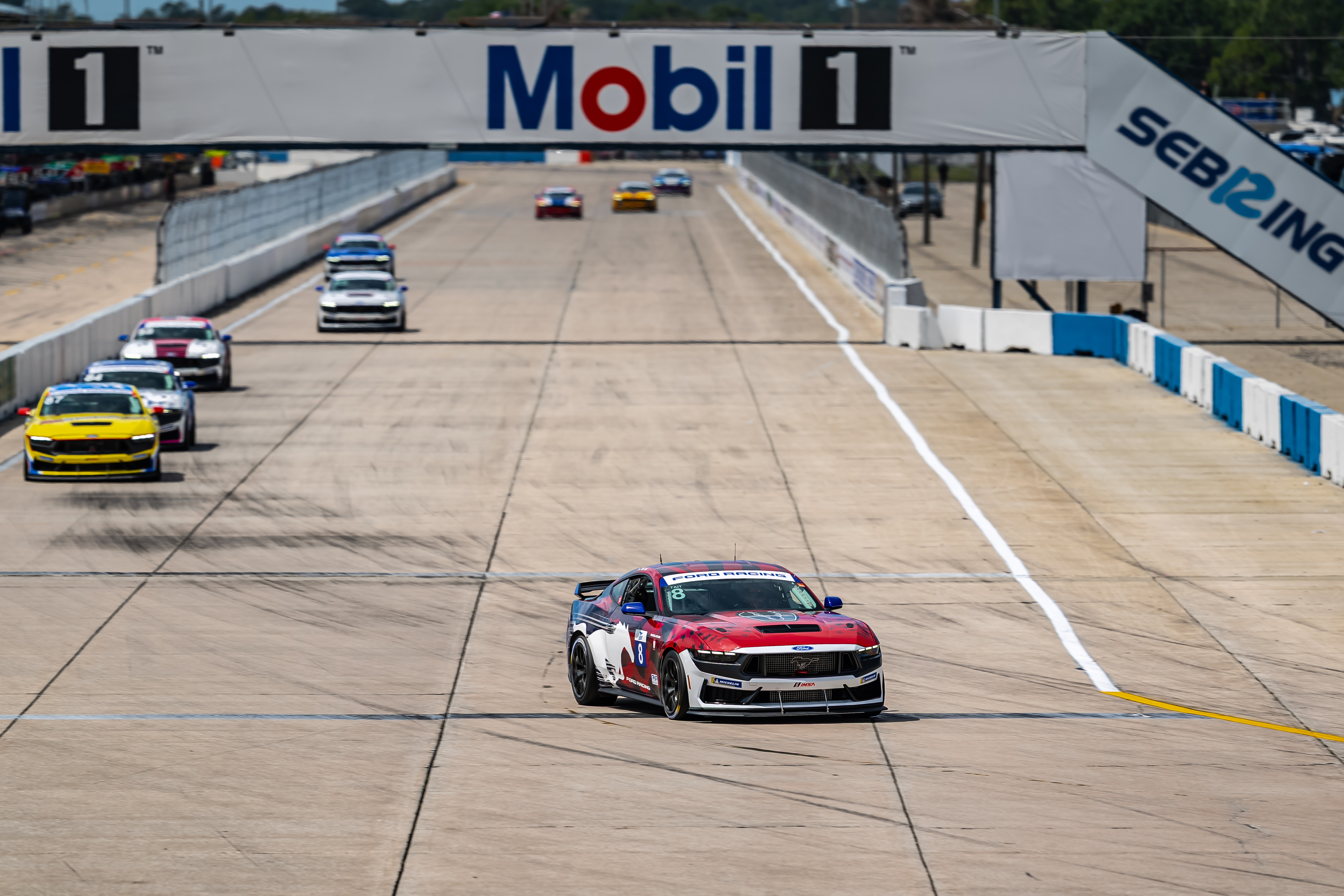 Red and white Ford Mustang race car in front of a pack of other race cars on a race track under a Mobil 1 banner.