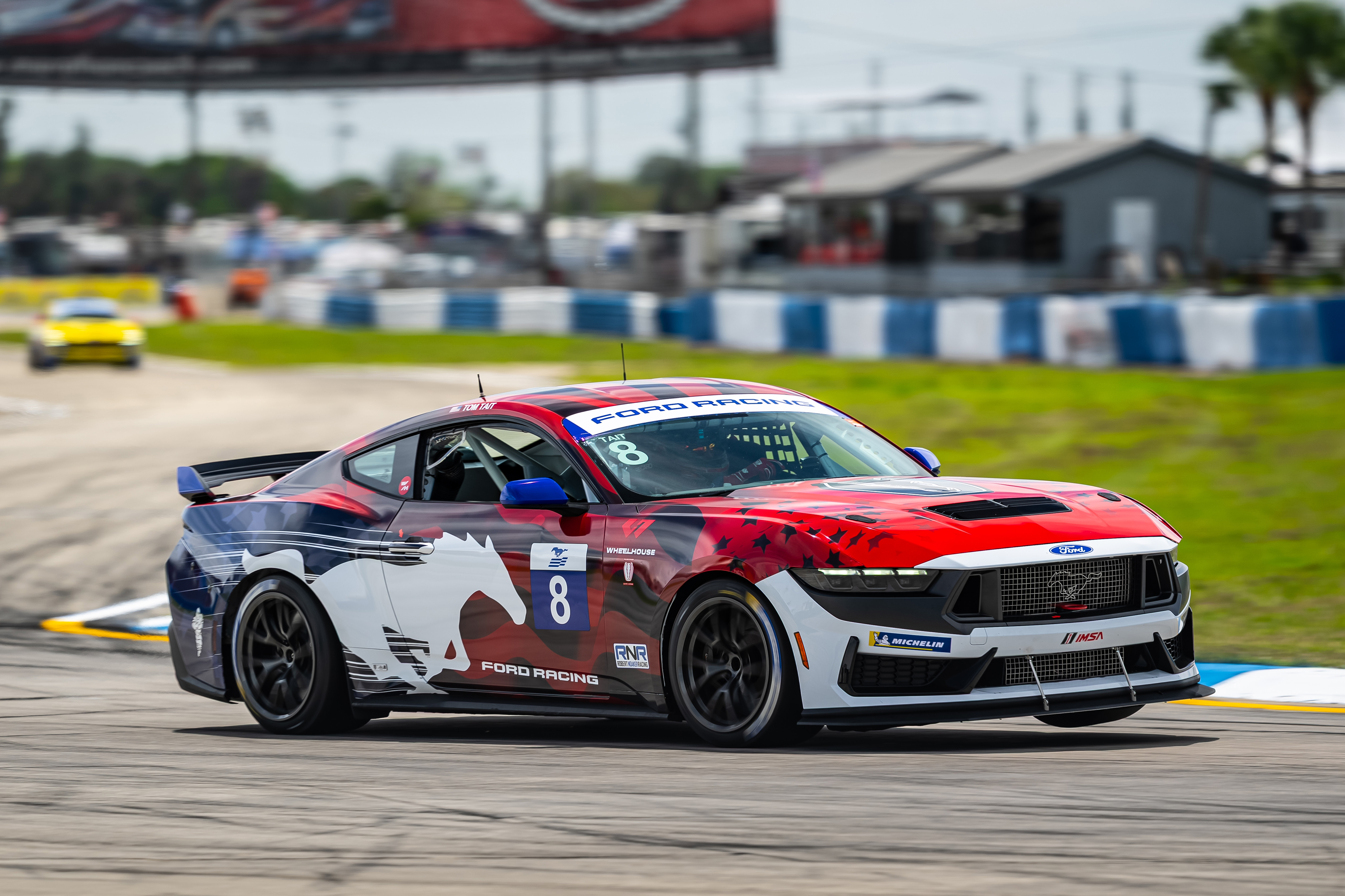 Red, white, and blue Ford Mustang race car with number 8 and Mustang logo racing on track.
