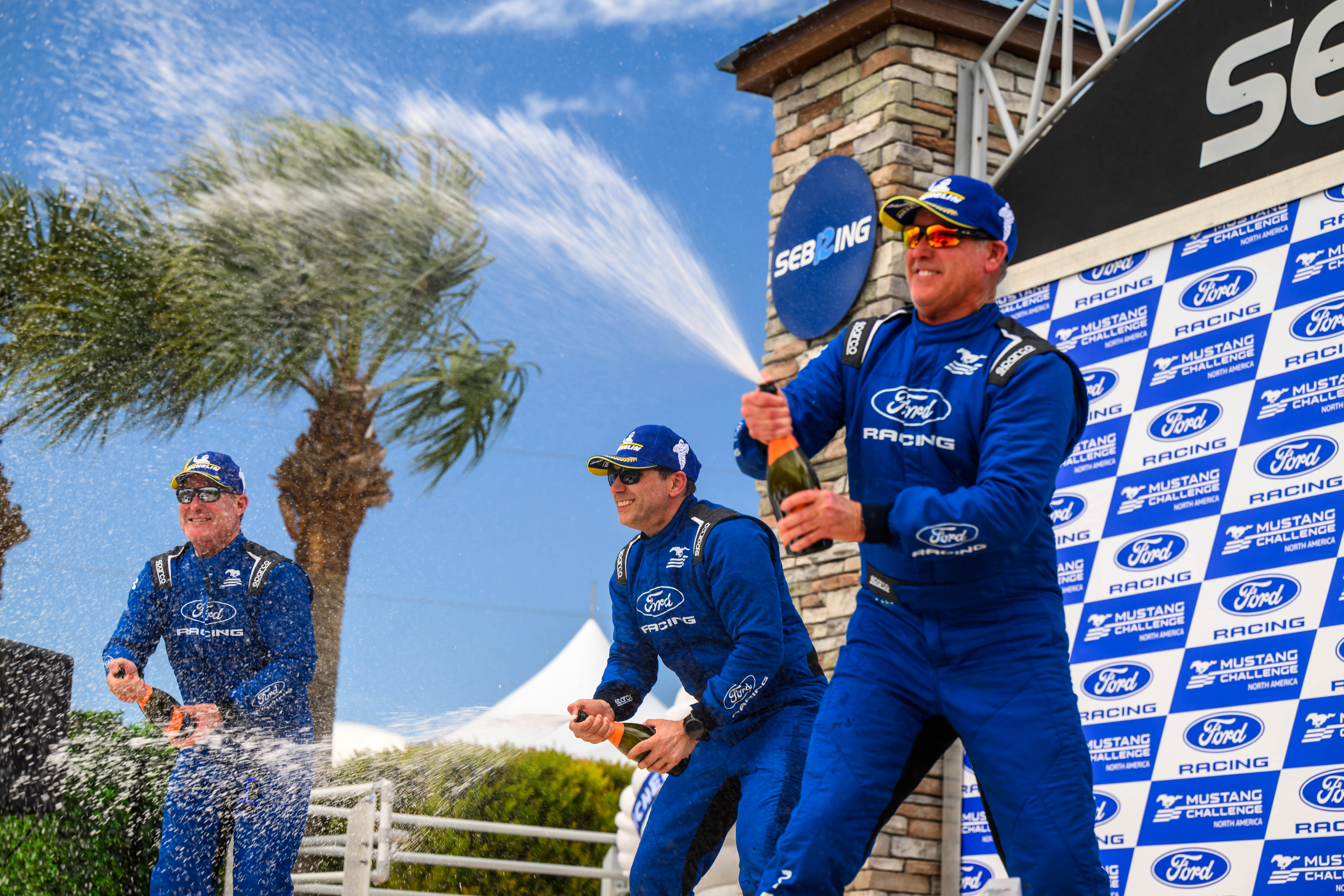 Three men in blue Ford Racing suits celebrating by spraying champagne at Sebring race event podium.
