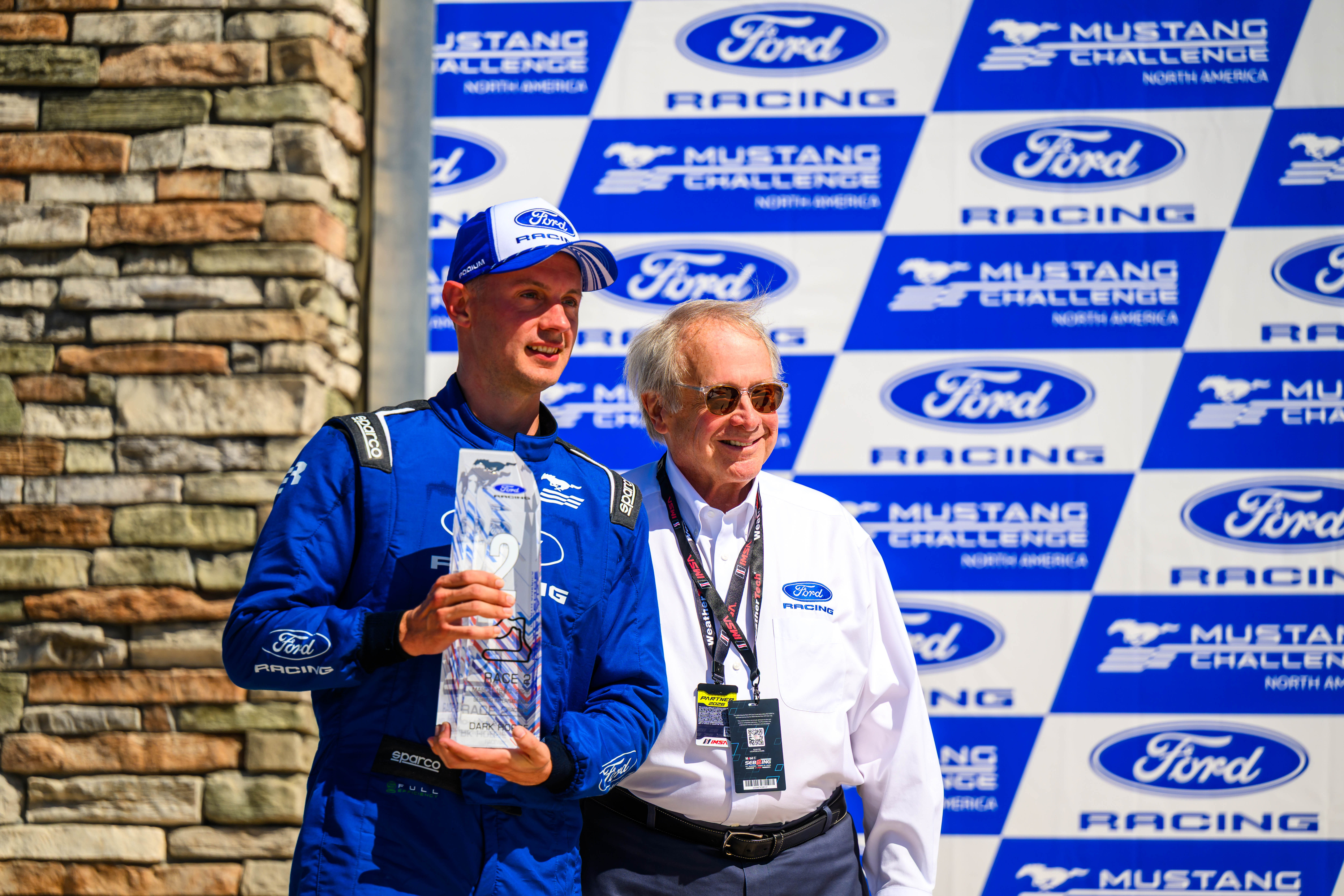 Race car driver in blue Ford Racing suit holding a trophy next to a man in white Ford Racing shirt in front of Ford Racing and Mustang Challenge backdrop.