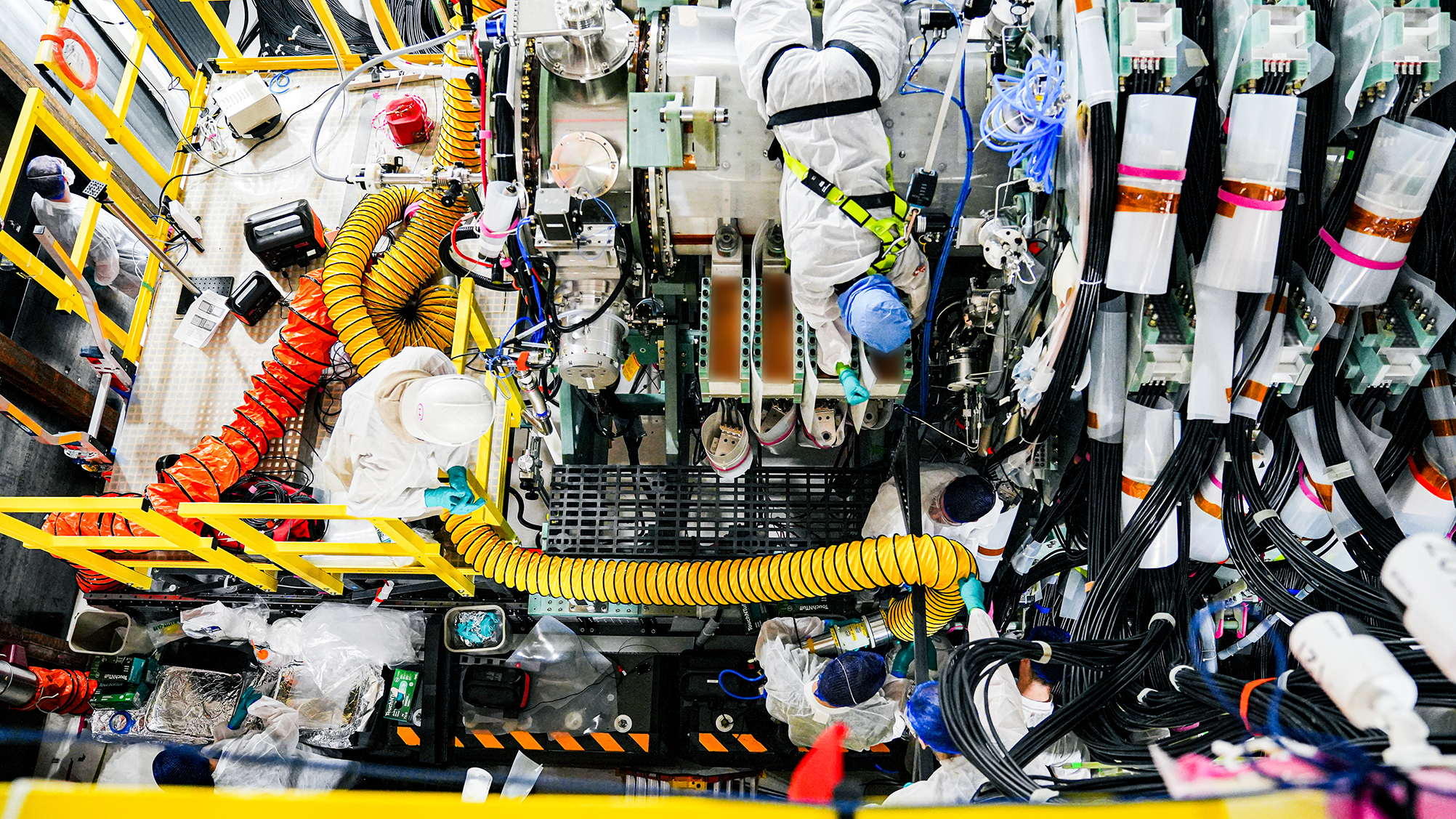 Technicians in protective suits and helmets working on complex machinery with cables and ventilation ducts in an industrial setting.