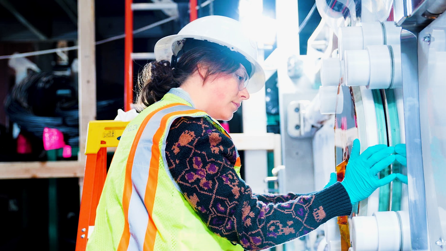 Worker wearing a hard hat, safety vest, and gloves operating industrial machinery.