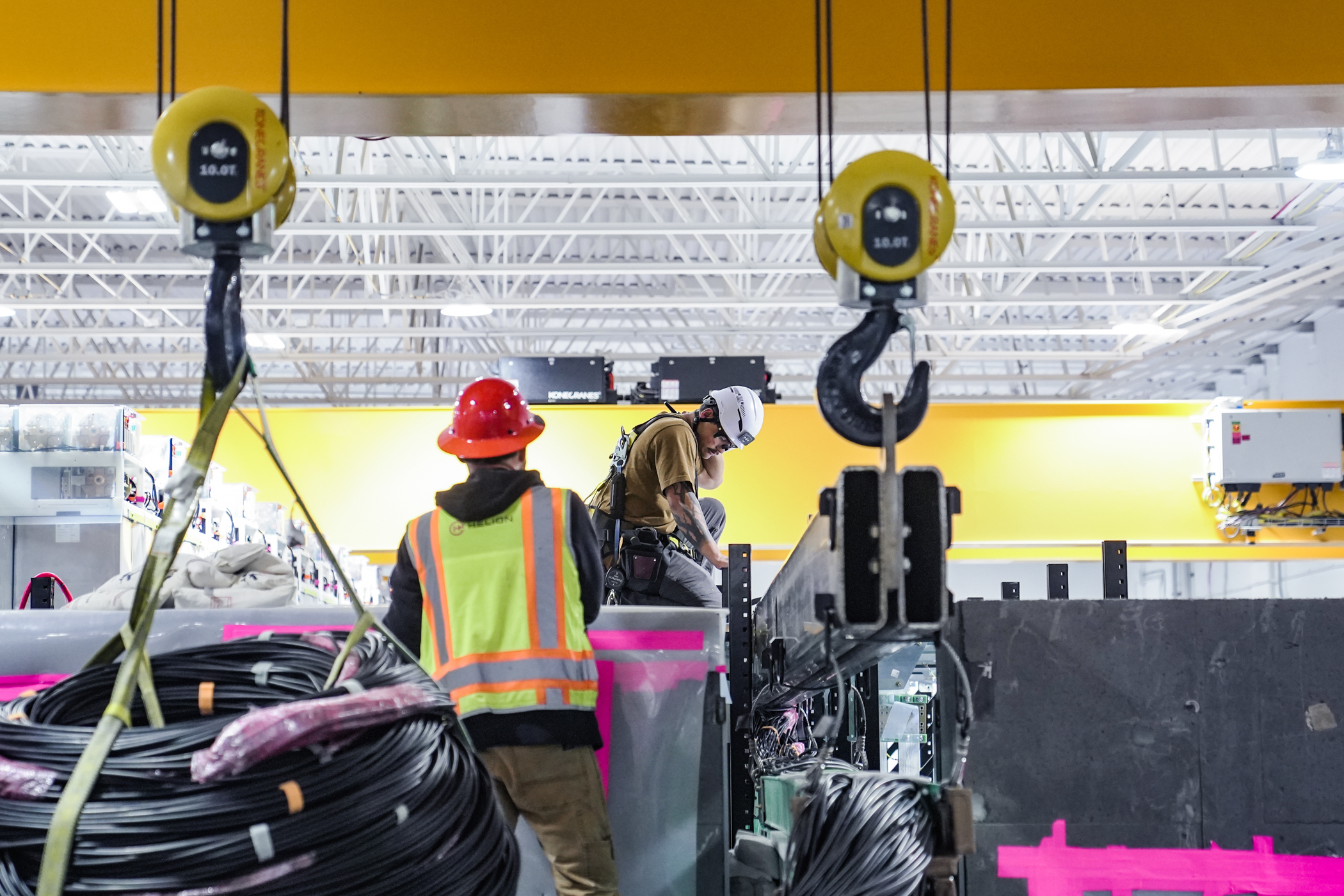 Two workers wearing safety helmets and vests in an industrial setting with heavy cables and lifting equipment.