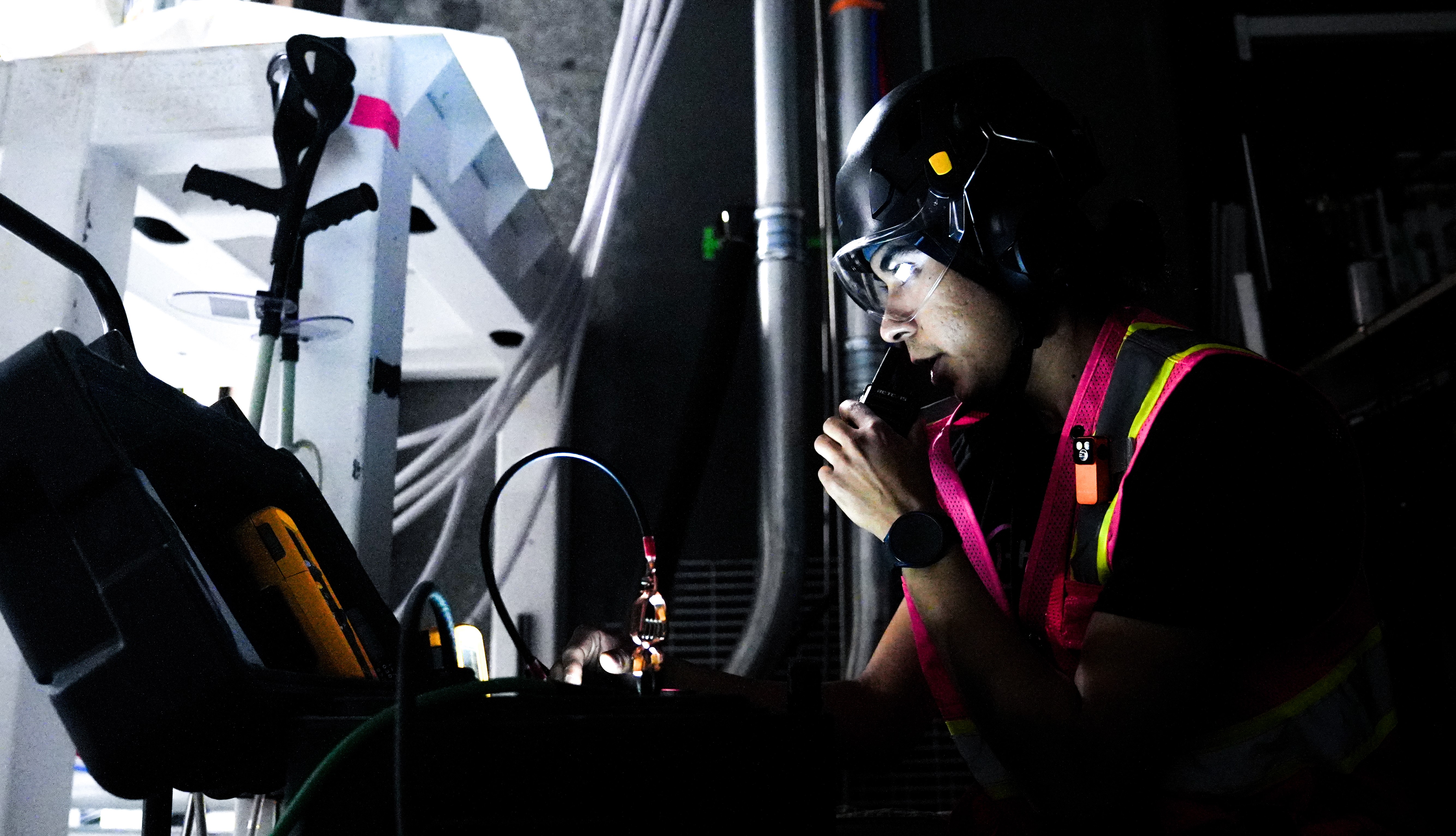Technician in safety vest and helmet speaking into a radio while working with connected cables on industrial equipment in a dimly lit environment.