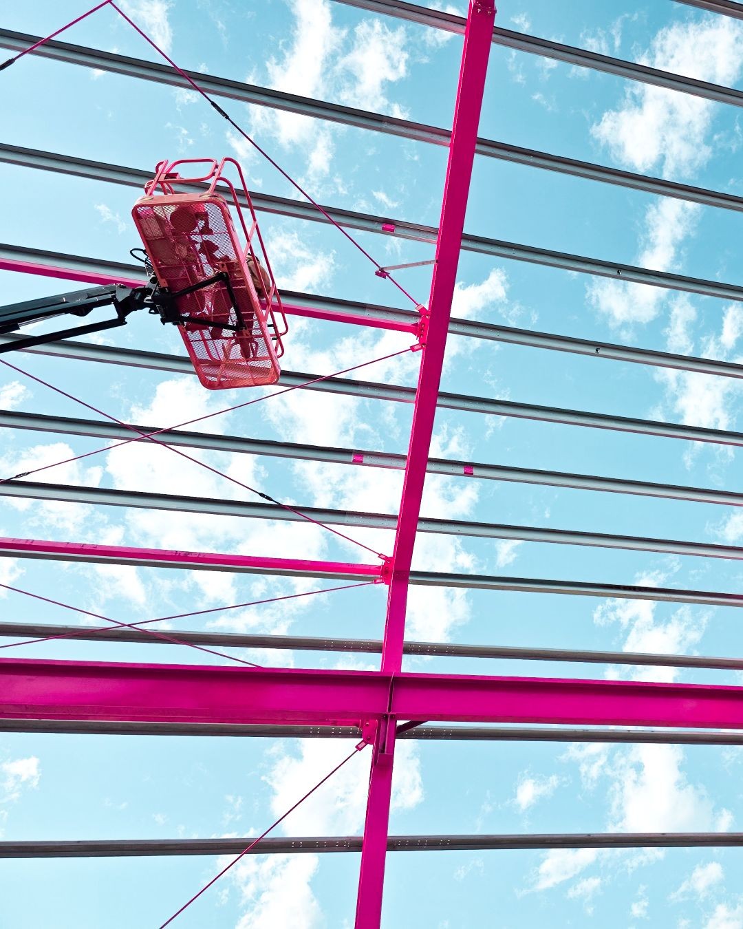 Construction lift platform elevated near pink and grey steel beams under a partly cloudy blue sky.