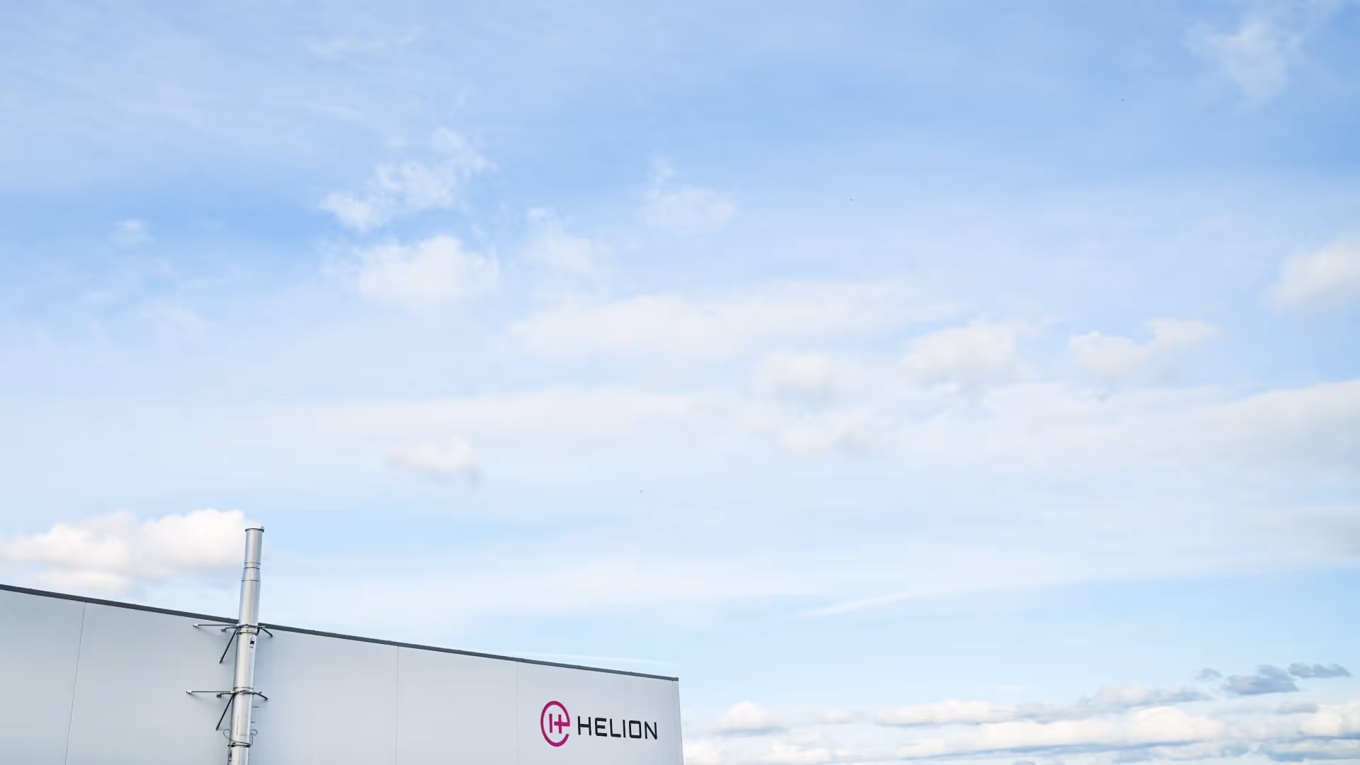 Exterior of a light gray building with a vertical exhaust pipe and the Helion logo on the wall against a partly cloudy blue sky.