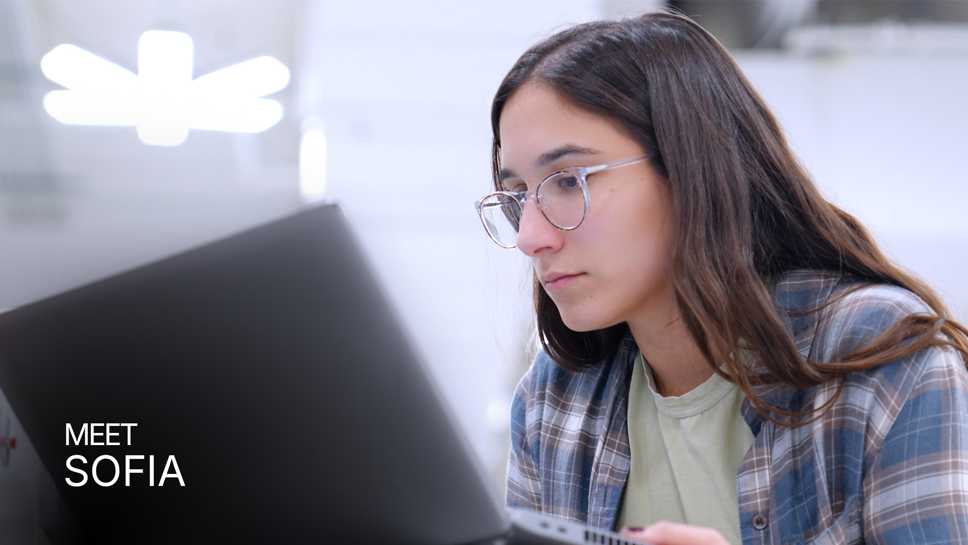 Young woman with glasses focused on her laptop in a bright indoor setting.