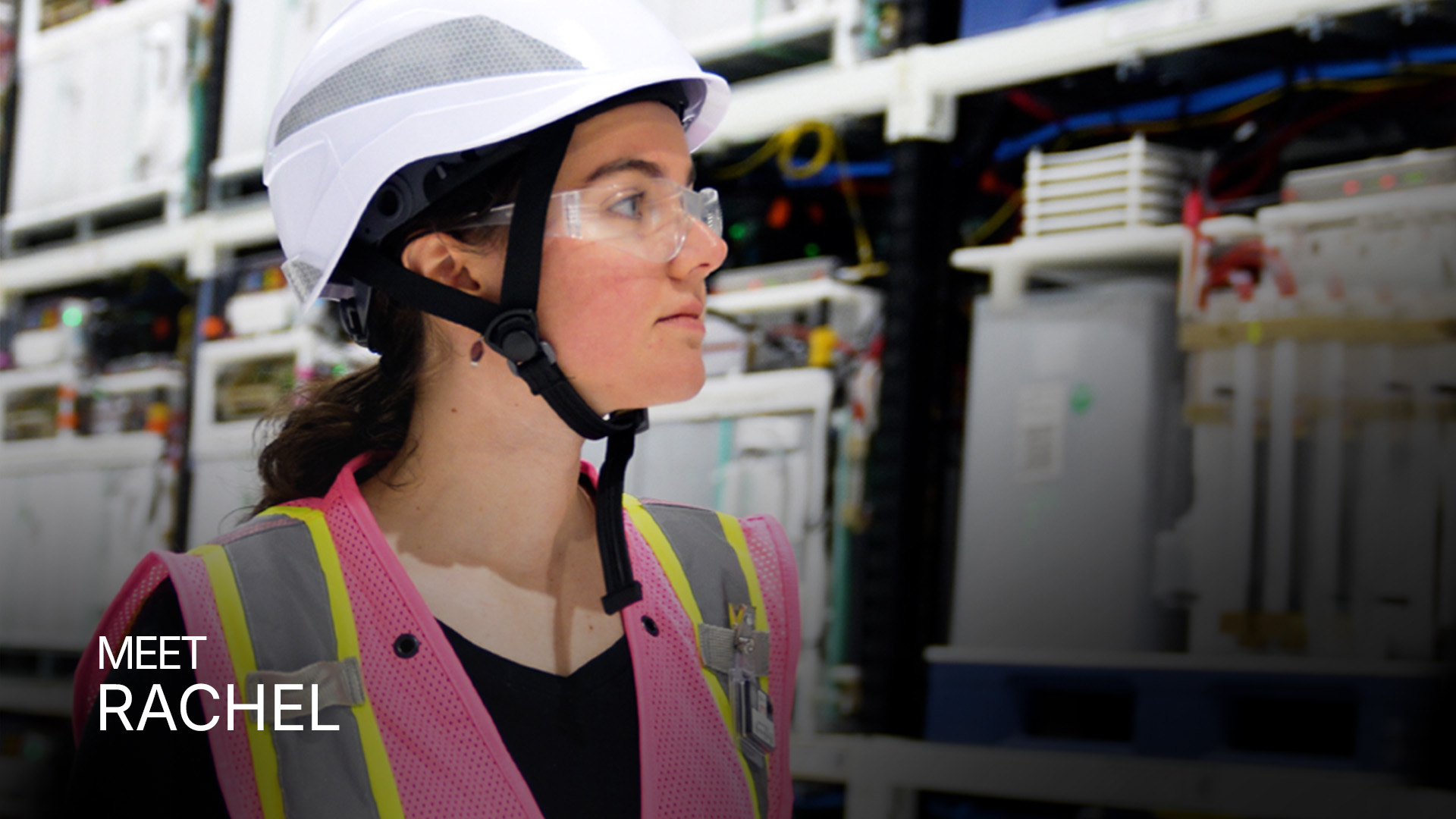 Woman wearing a white hard hat, safety glasses, and a pink reflective safety vest in an industrial setting.