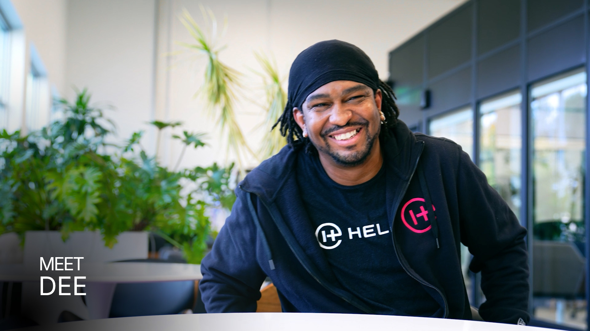 Smiling man wearing a black Helion-branded hoodie sitting at a table in a modern office with plants in the background.
