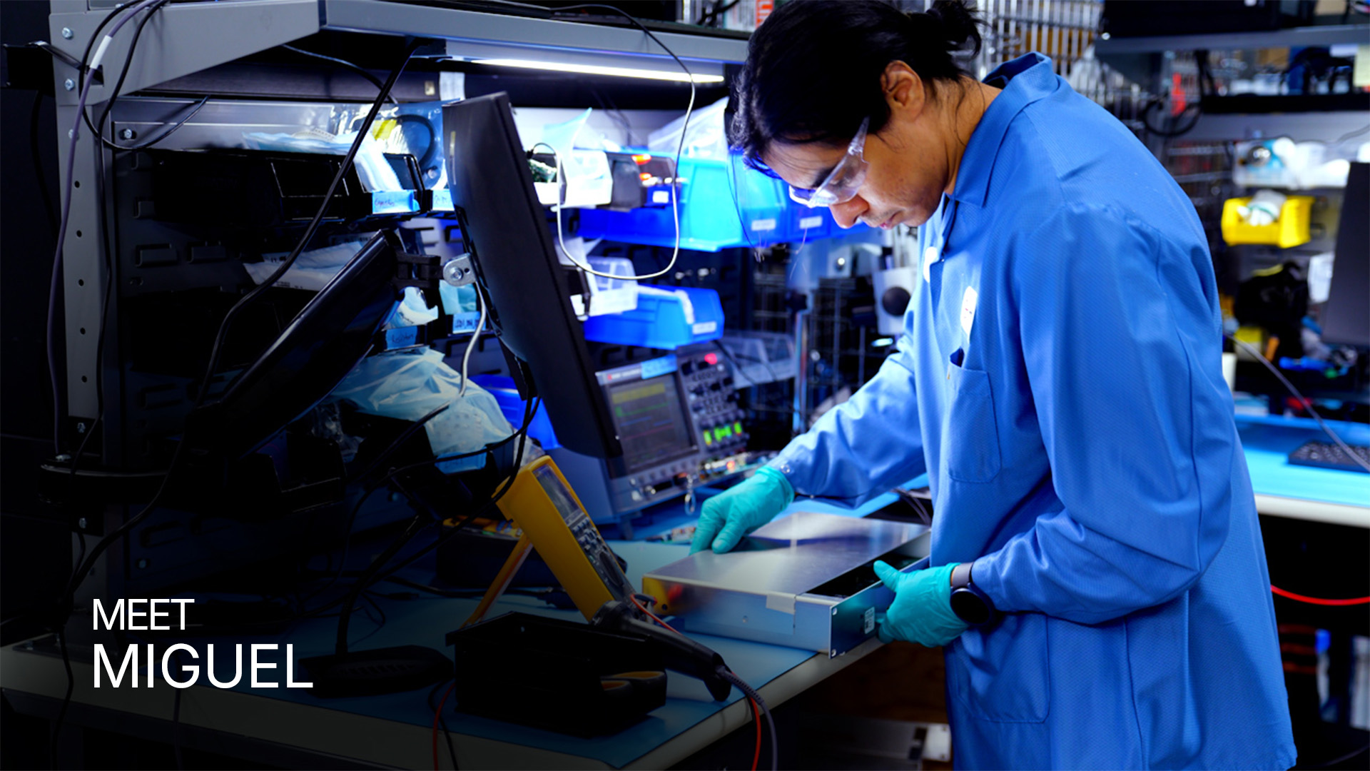 Technician in blue lab coat and gloves working on electronic equipment at a lab bench.