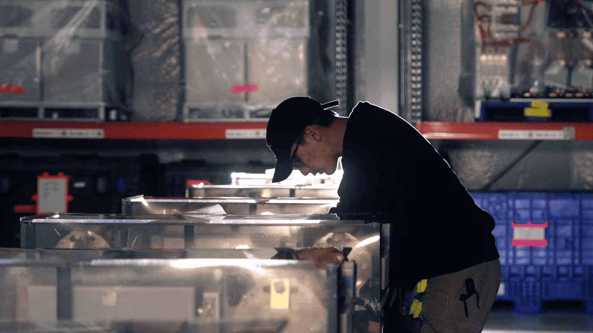 Man wearing a black cap and glasses inspecting equipment in an industrial setting.