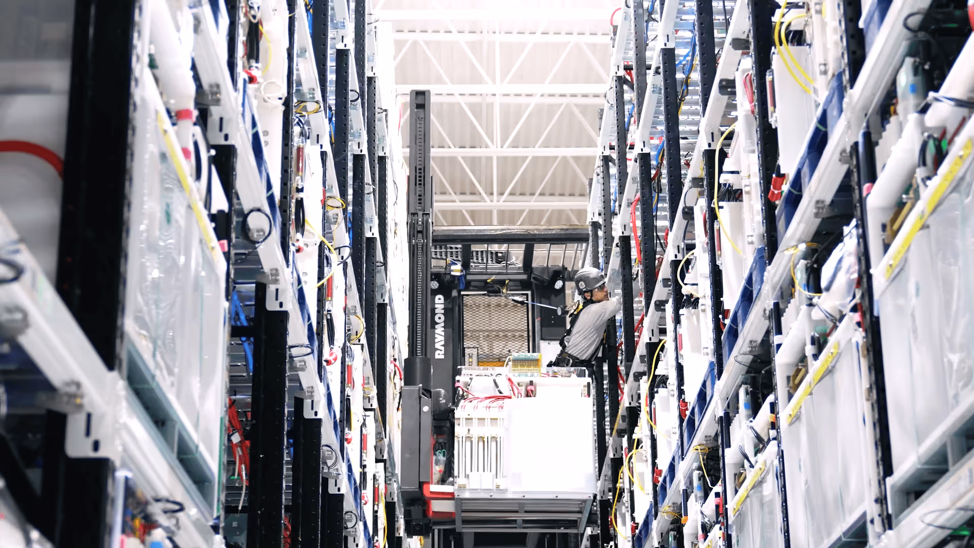 Technician wearing a helmet operating a forklift between tall racking shelves filled with capacitors.