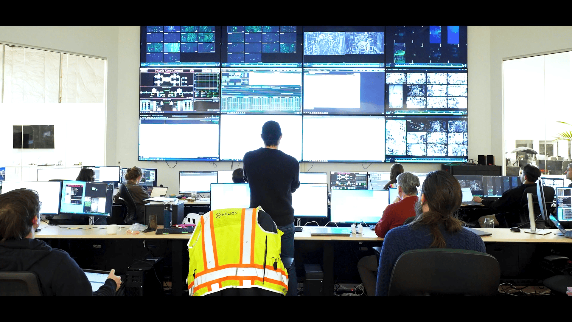 Office with multiple employees at desks, large wall of monitors displaying data and surveillance feeds.