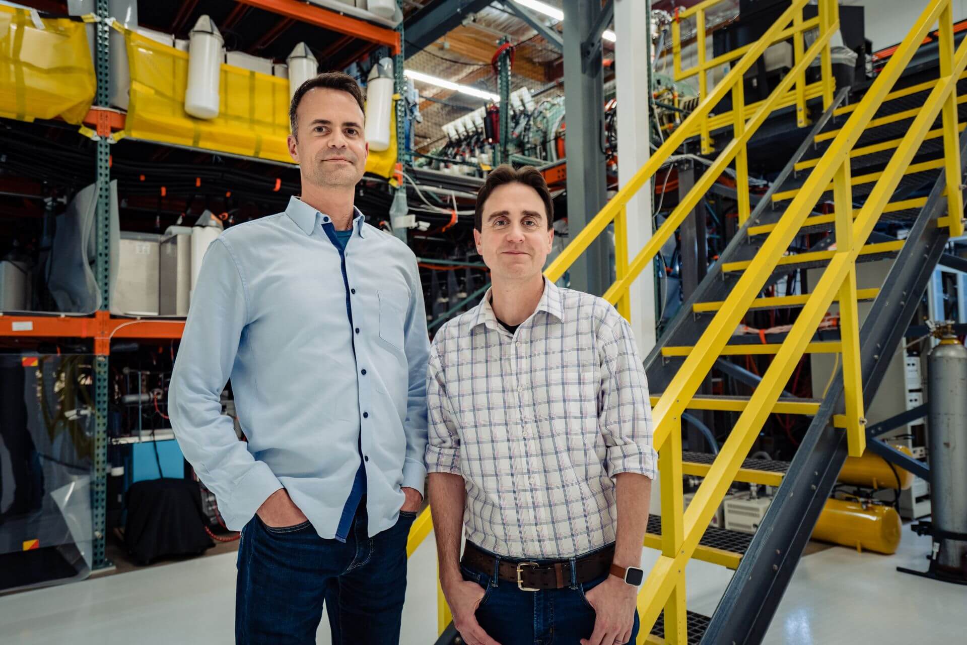 Chris Pihl and David Kirtley standing in an industrial setting with machinery and a yellow stairway behind them.