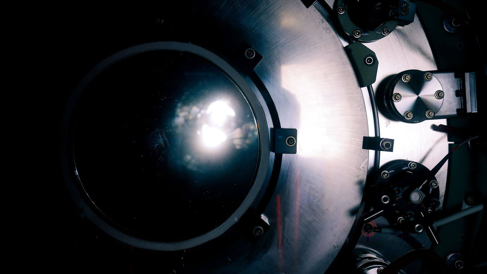 Close-up view of a large circular metallic machine part with bolts and a transparent central window reflecting light.
