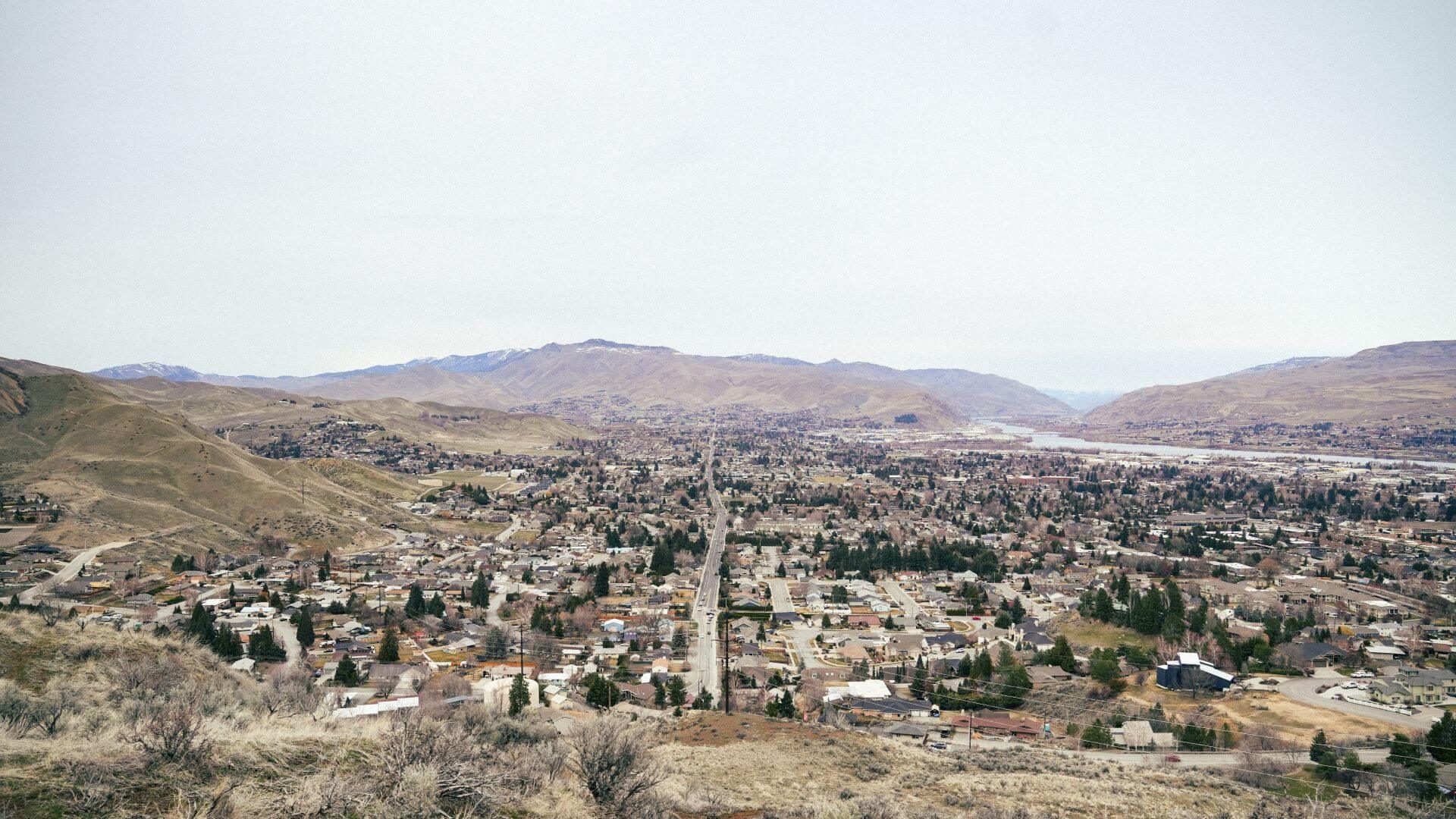 A wide aerial view of Wenatchee City with a road running through residential neighborhoods, surrounded by dry hills and distant mountains.