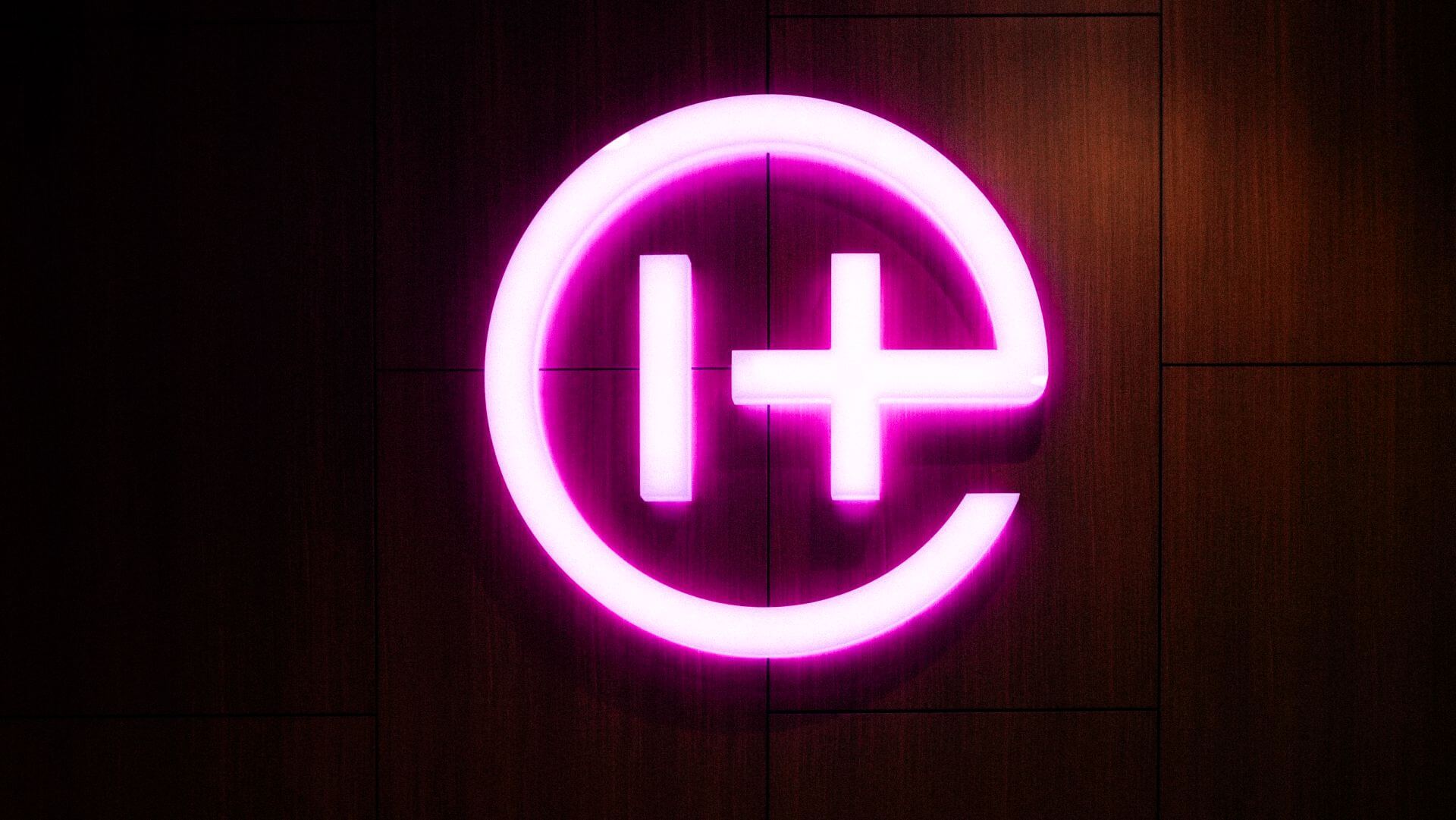 Bright pink neon sign of the Helion logo, a stylized 'H' inside a circle on a dark wooden wall.