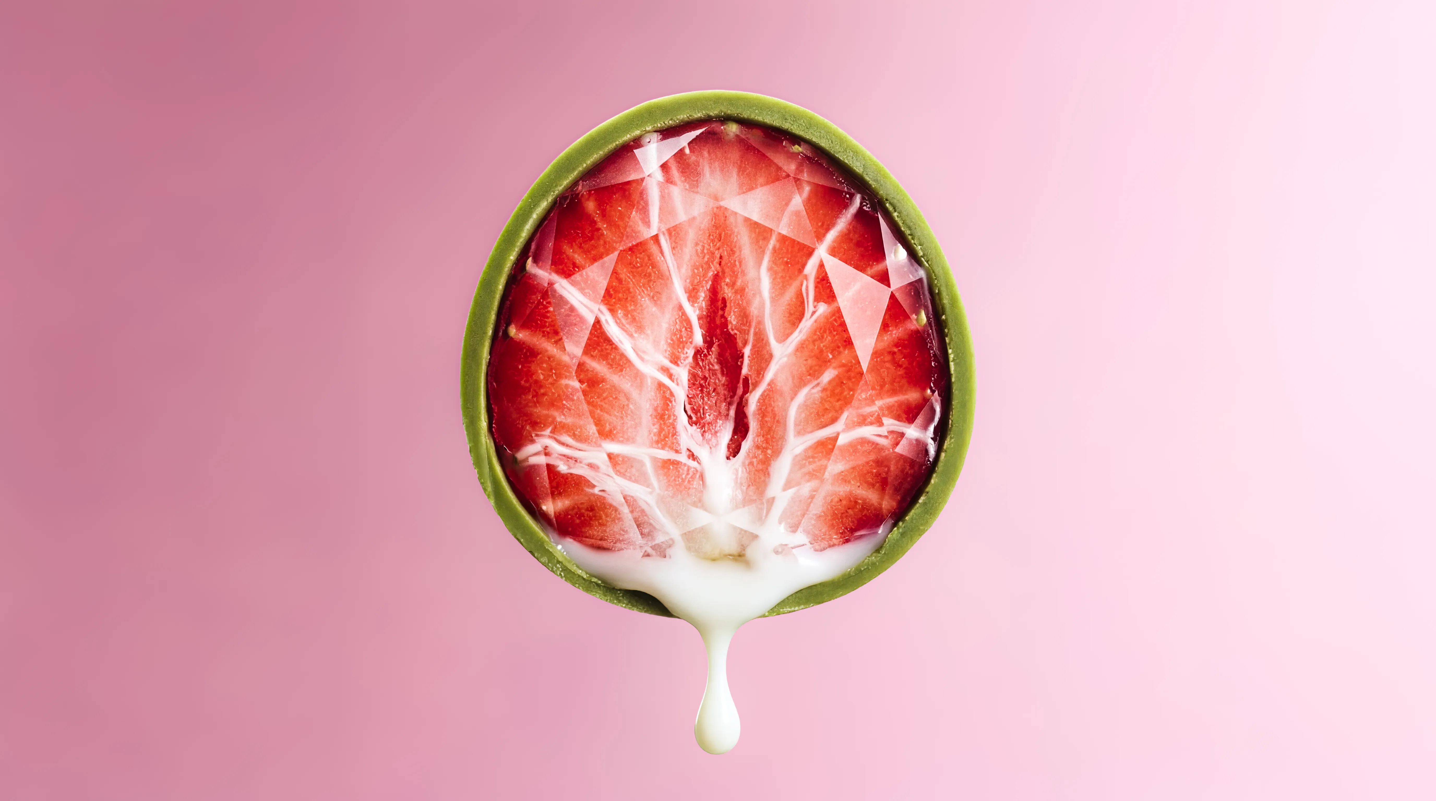 Close-up of a halved strawberry held by a hand coated in green, dripping white liquid on a pink background.