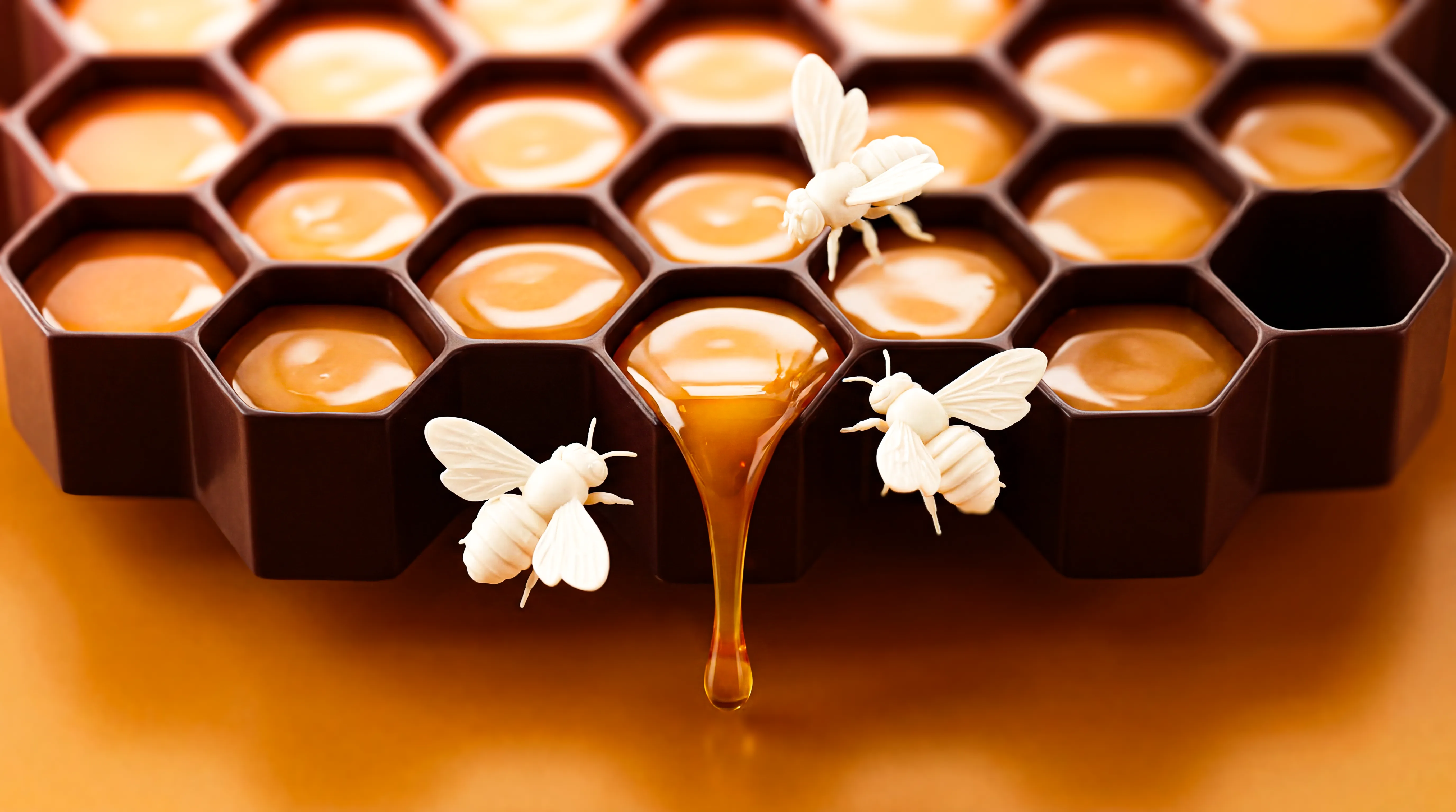 Honeycomb-shaped tray filled with golden honey, with three white bee figurines on and around it and honey dripping over one edge.