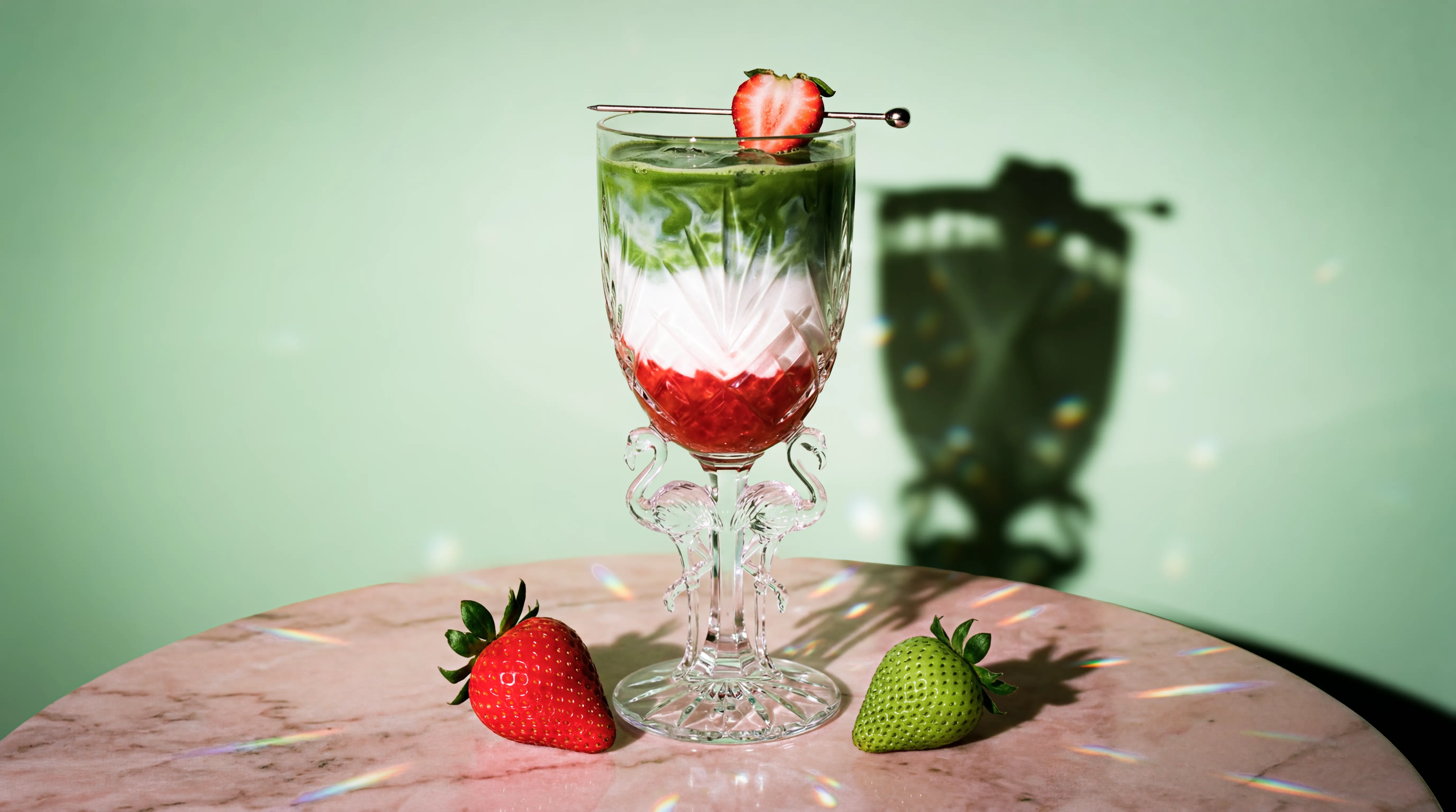 Layered iced drink in a decorative glass with flamingo-shaped stem, garnished with a strawberry slice, on a marble table with a green strawberry and a red strawberry beside it.