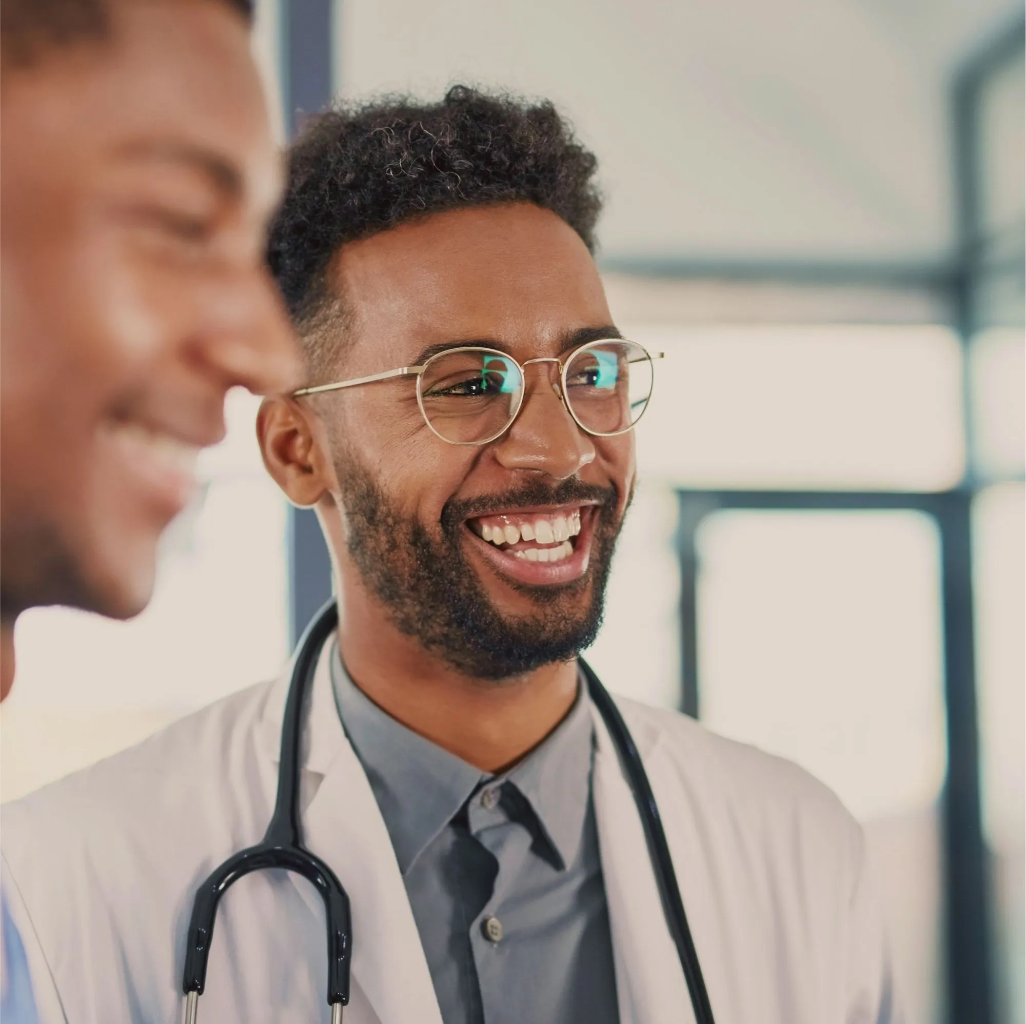 Smiling male doctor wearing glasses and a stethoscope around his neck in a bright medical office.