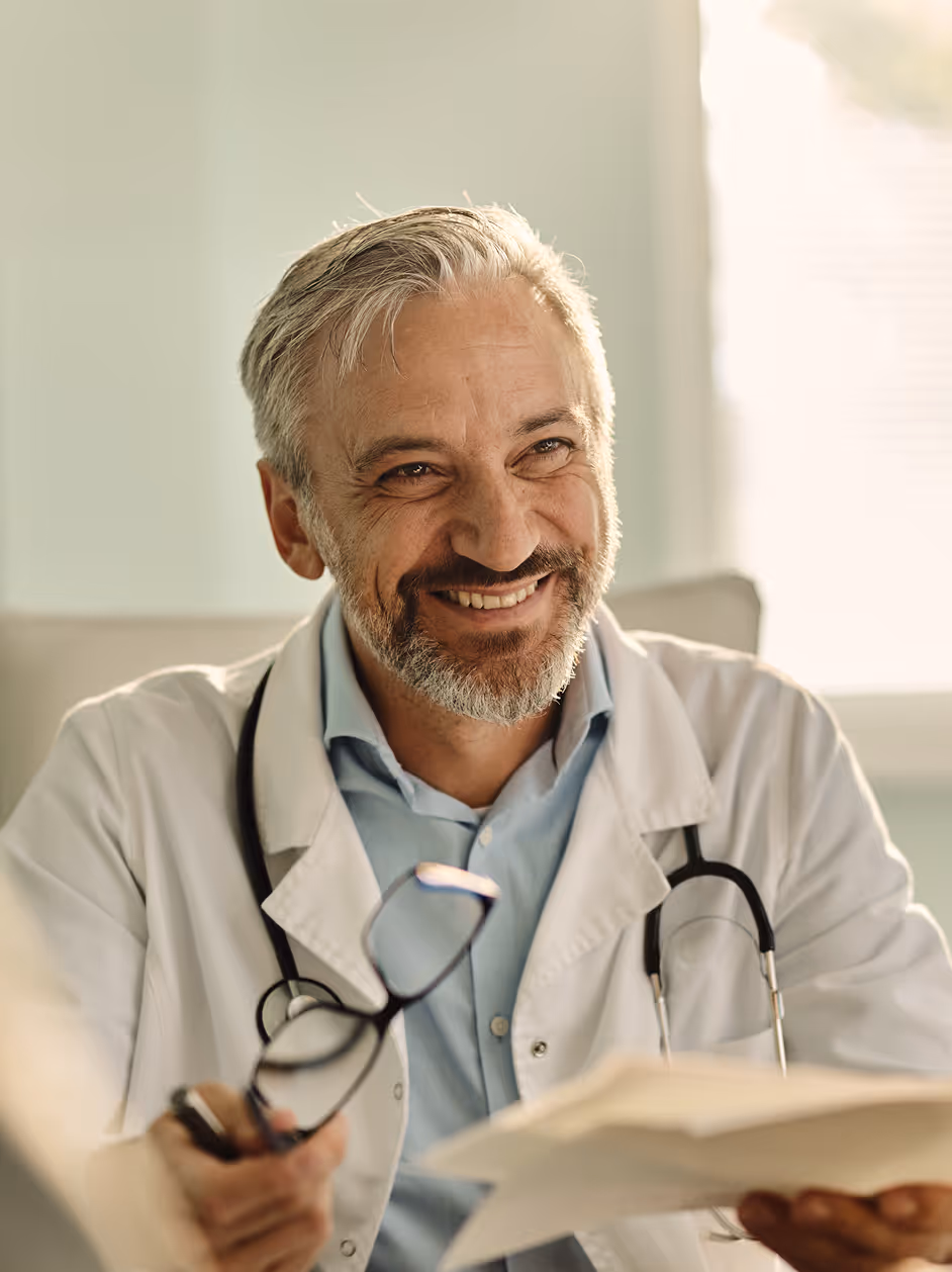 Smiling middle-aged male doctor in white coat holding glasses and papers in a bright office.