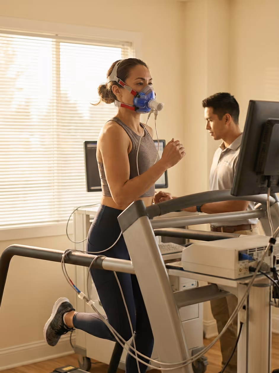 Woman running on a treadmill wearing a respiratory mask connected to medical equipment while a man monitors data on a computer.