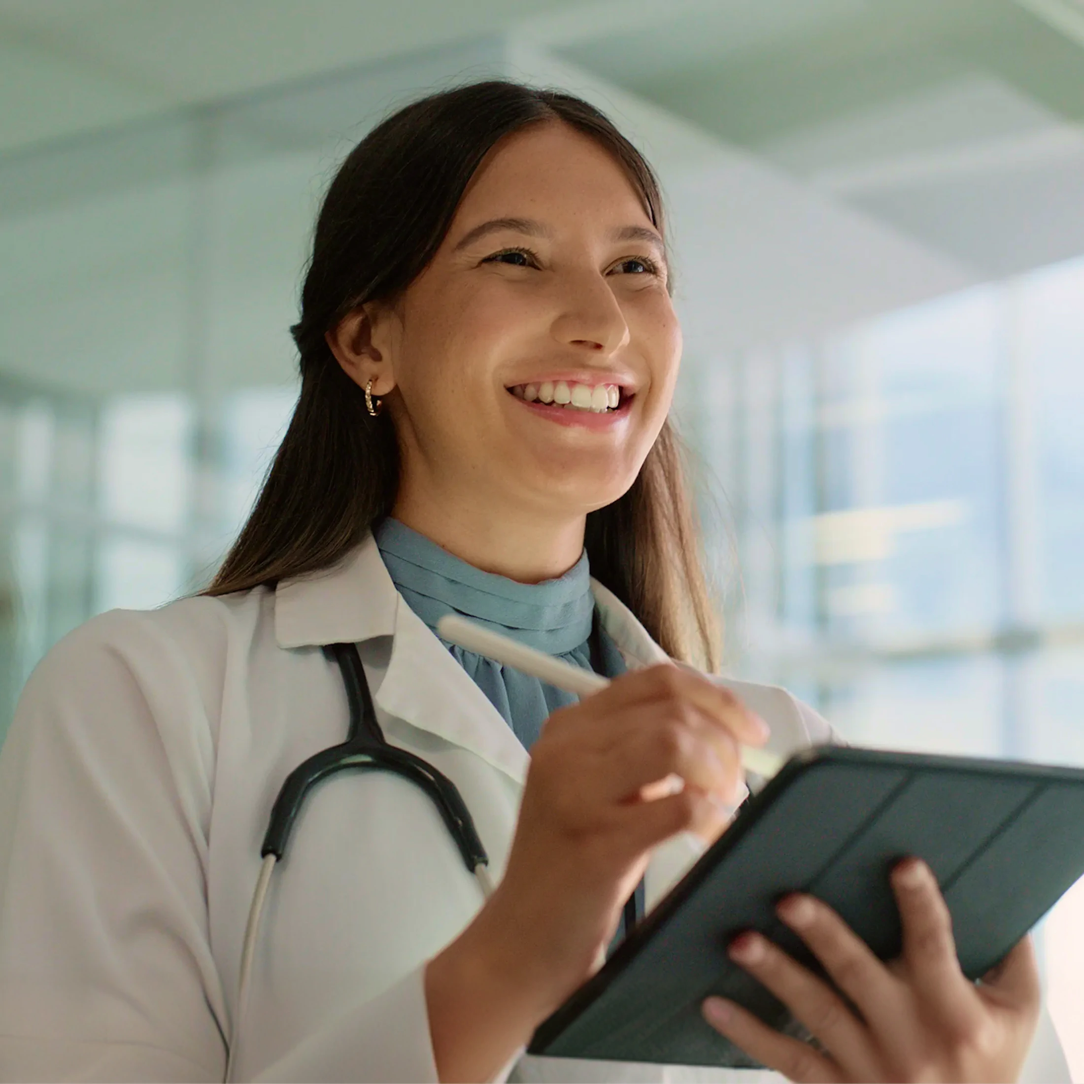 Smiling female doctor wearing a white coat and stethoscope, writing on a tablet with a stylus in a bright medical office.