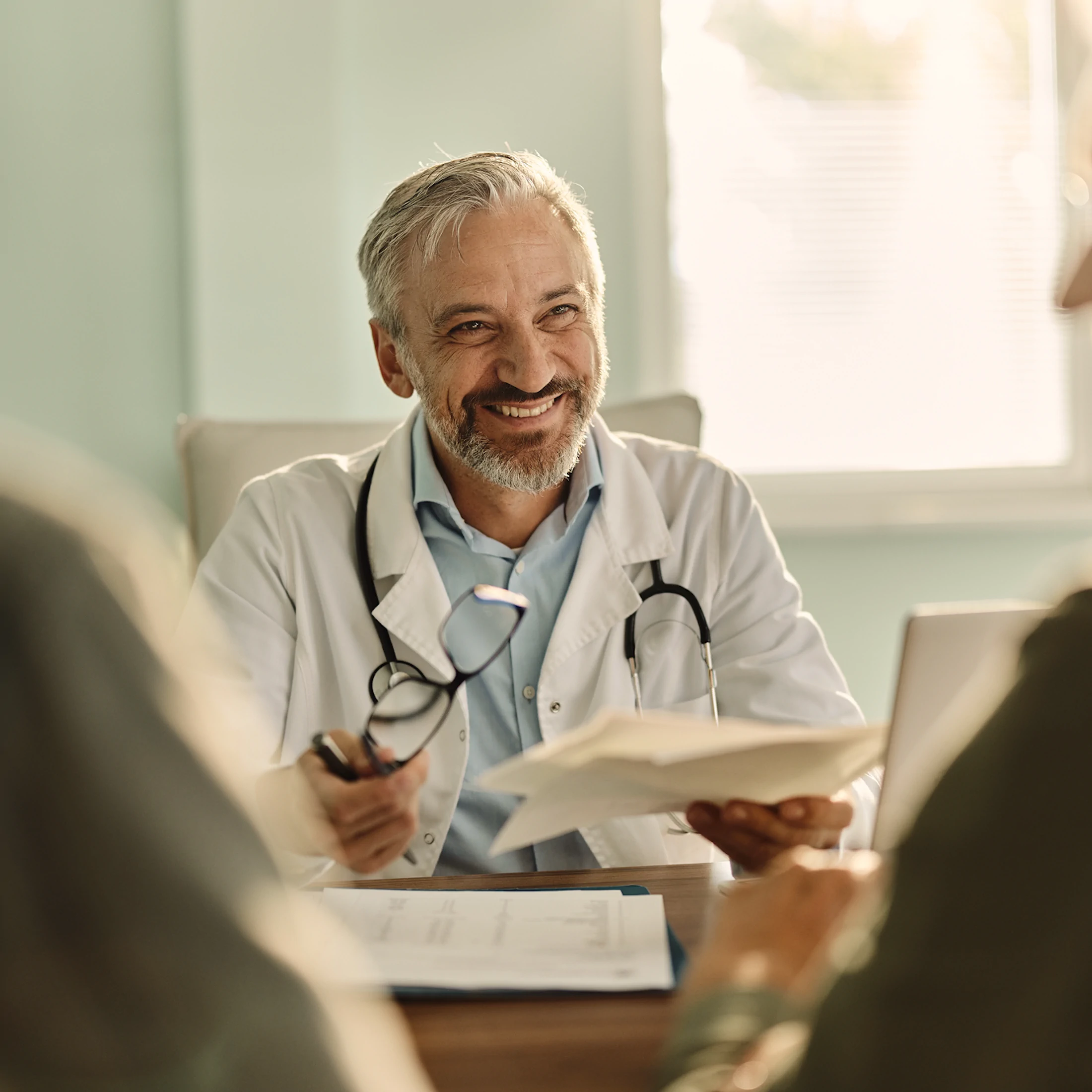 Smiling male doctor with gray hair and beard holding glasses and medical documents during consultation.
