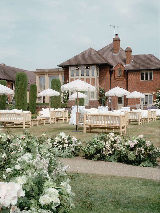 Outdoor wedding seating area with wooden benches, white umbrellas, and floral arrangements on a lawn.