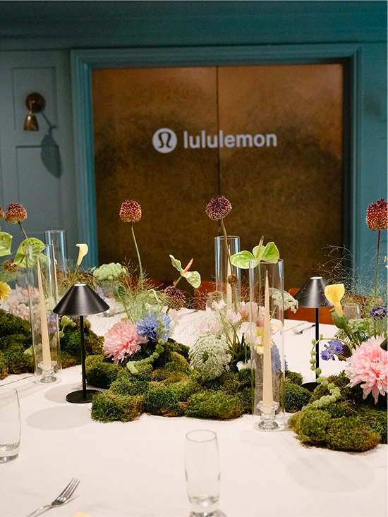 White table with floral centerpieces and candles in glass holders in front of a bronze door with lululemon logo.