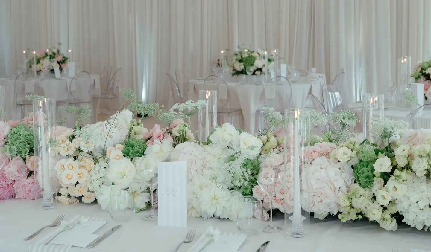 Elegant wedding table setting with white and pastel floral arrangements and lit glass candle holders.