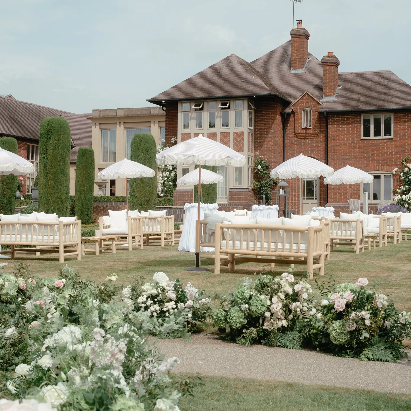 Outdoor seating area with wooden benches, white umbrellas, and floral arrangements in front of a brick house.