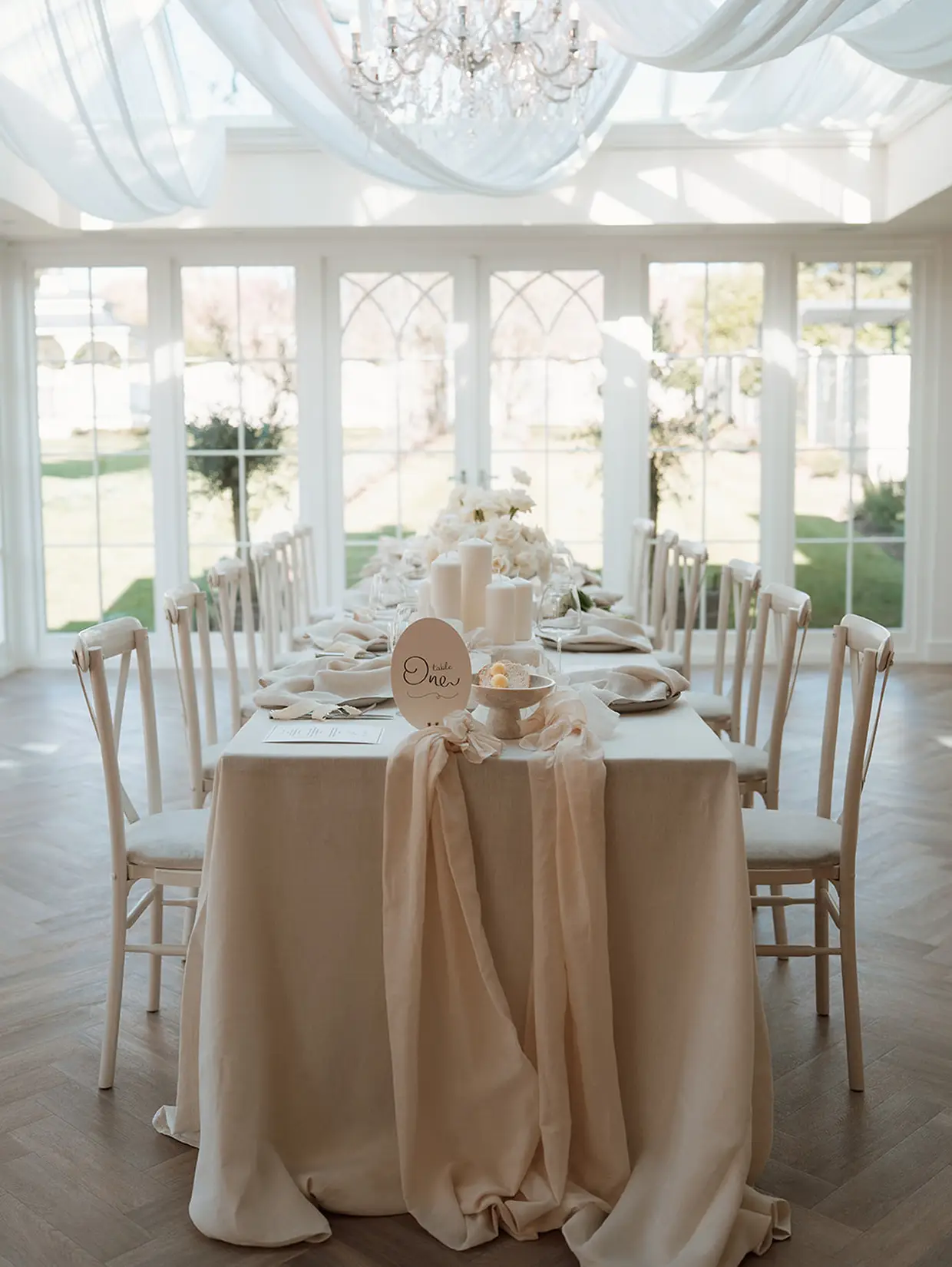 Elegant wedding table set with white linens, candles, flowers, and chairs in a bright room with large windows.