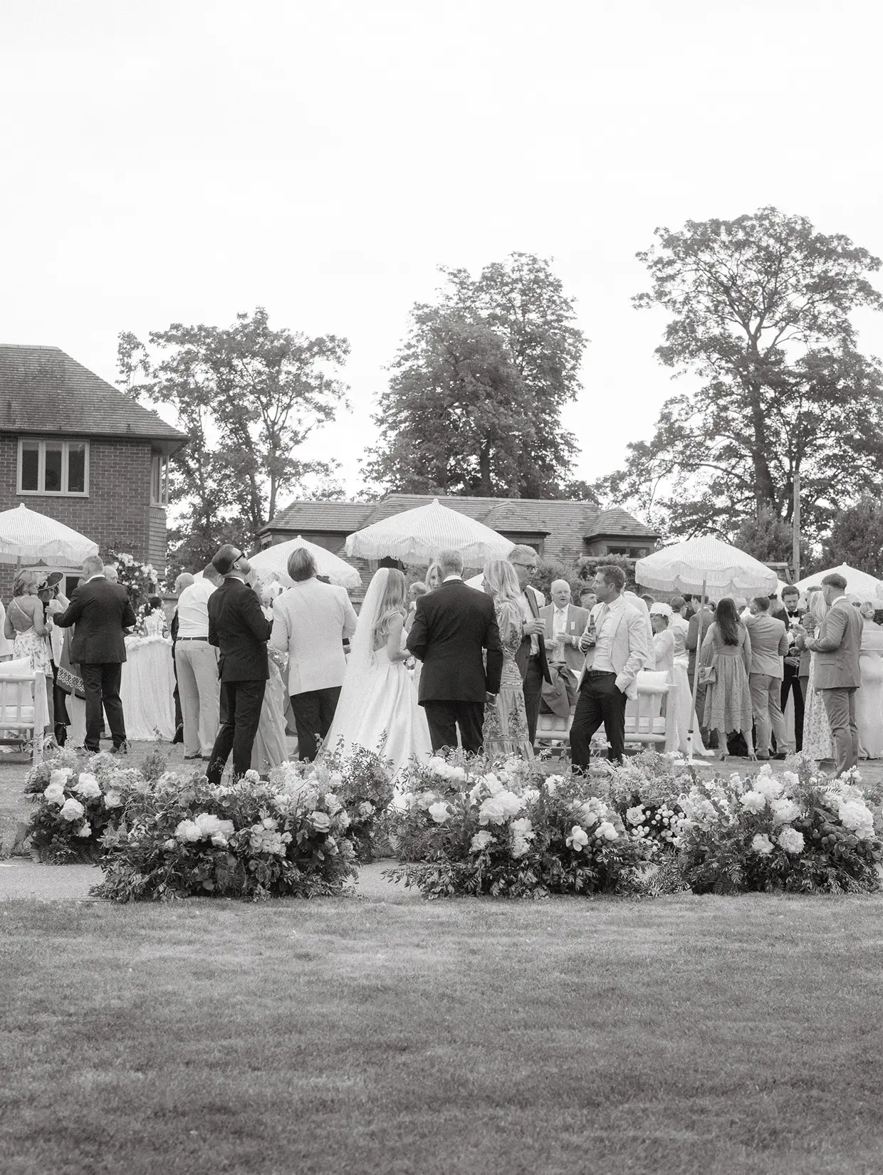 Outdoor wedding reception with guests mingling under umbrellas and floral arrangements in foreground.